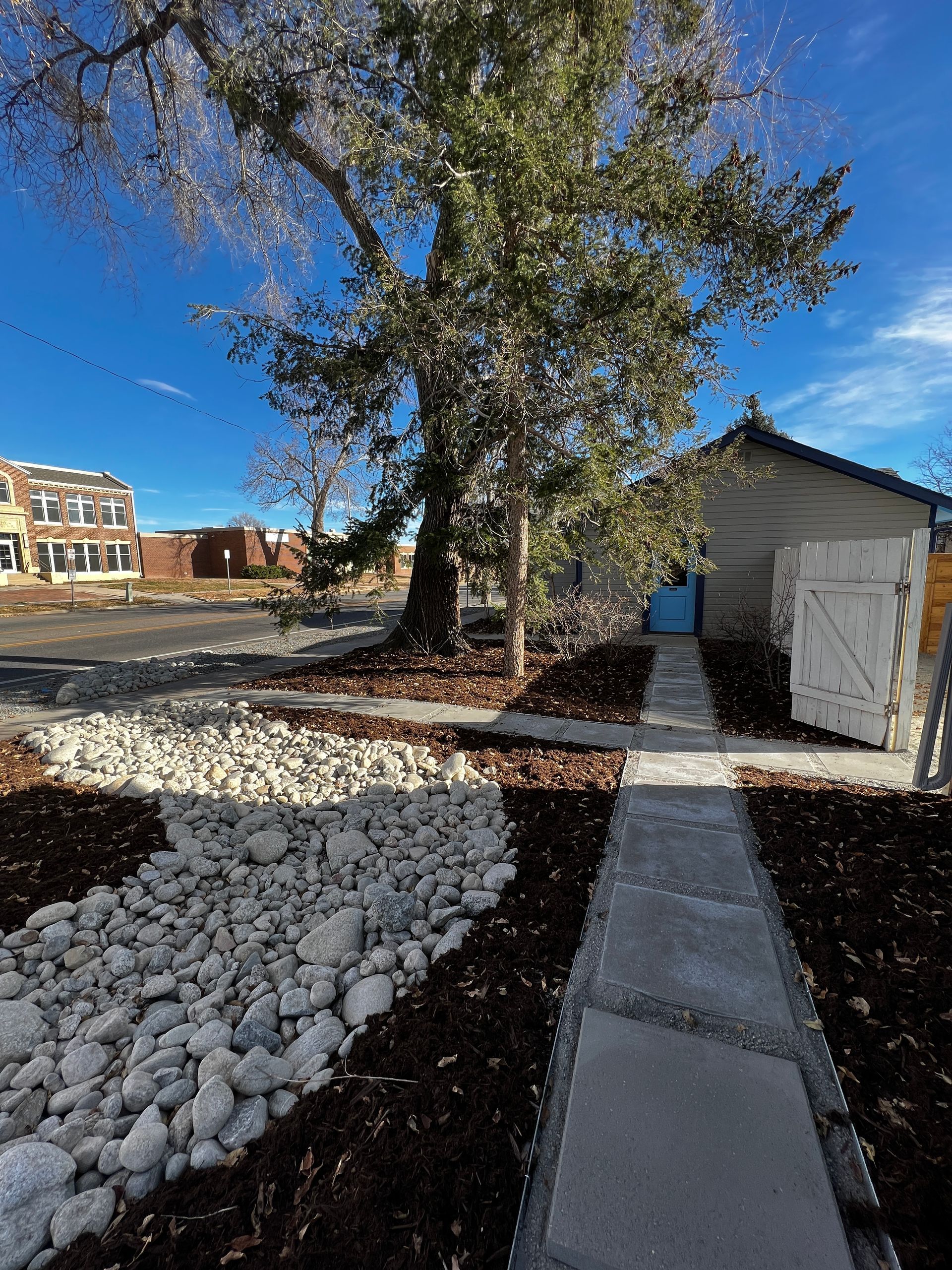 Stone path leads to small building with blue door, large tree overhead.