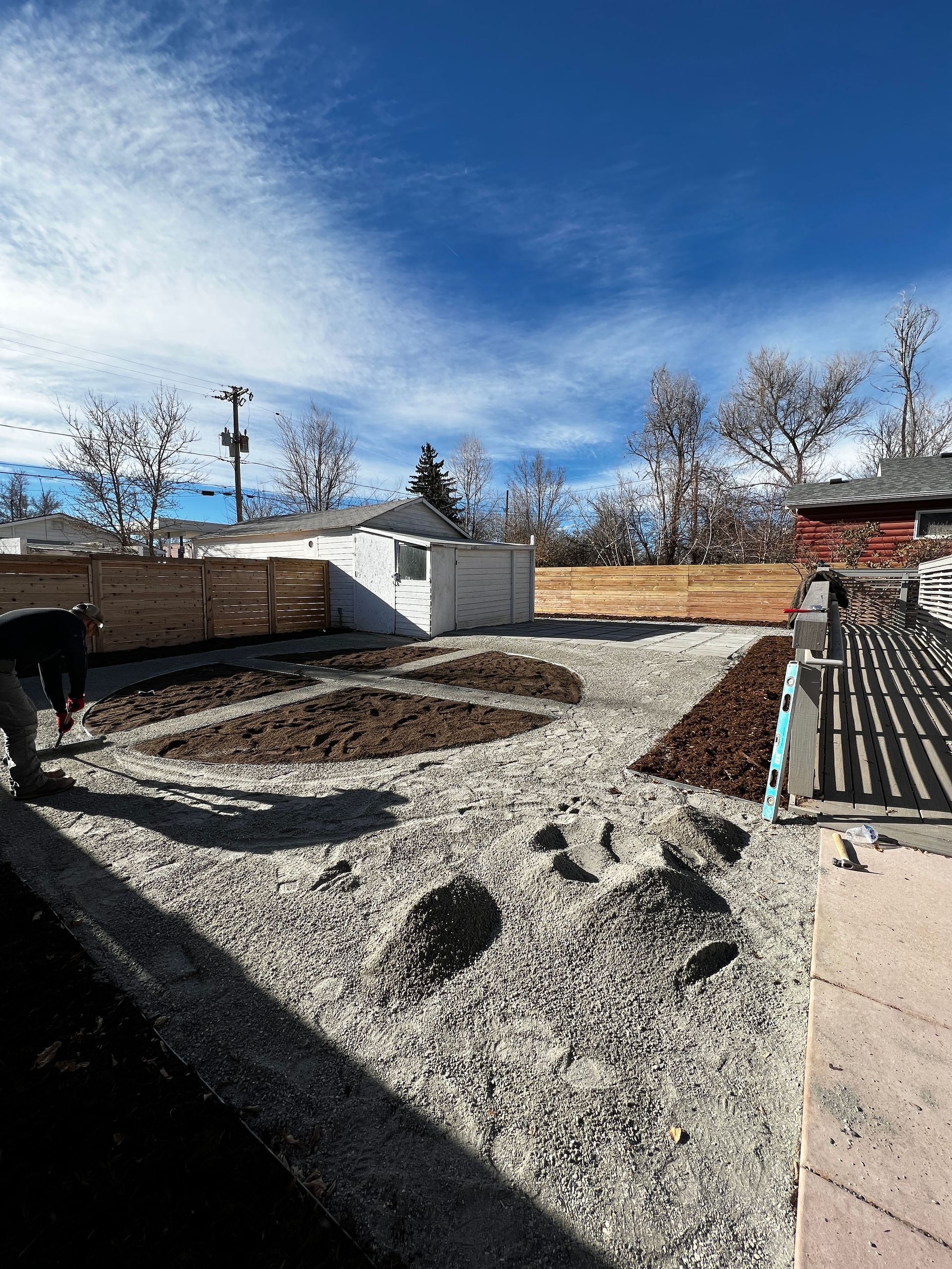 Backyard with gravel ground, wooden fence, and freshly tilled garden beds under a blue sky.