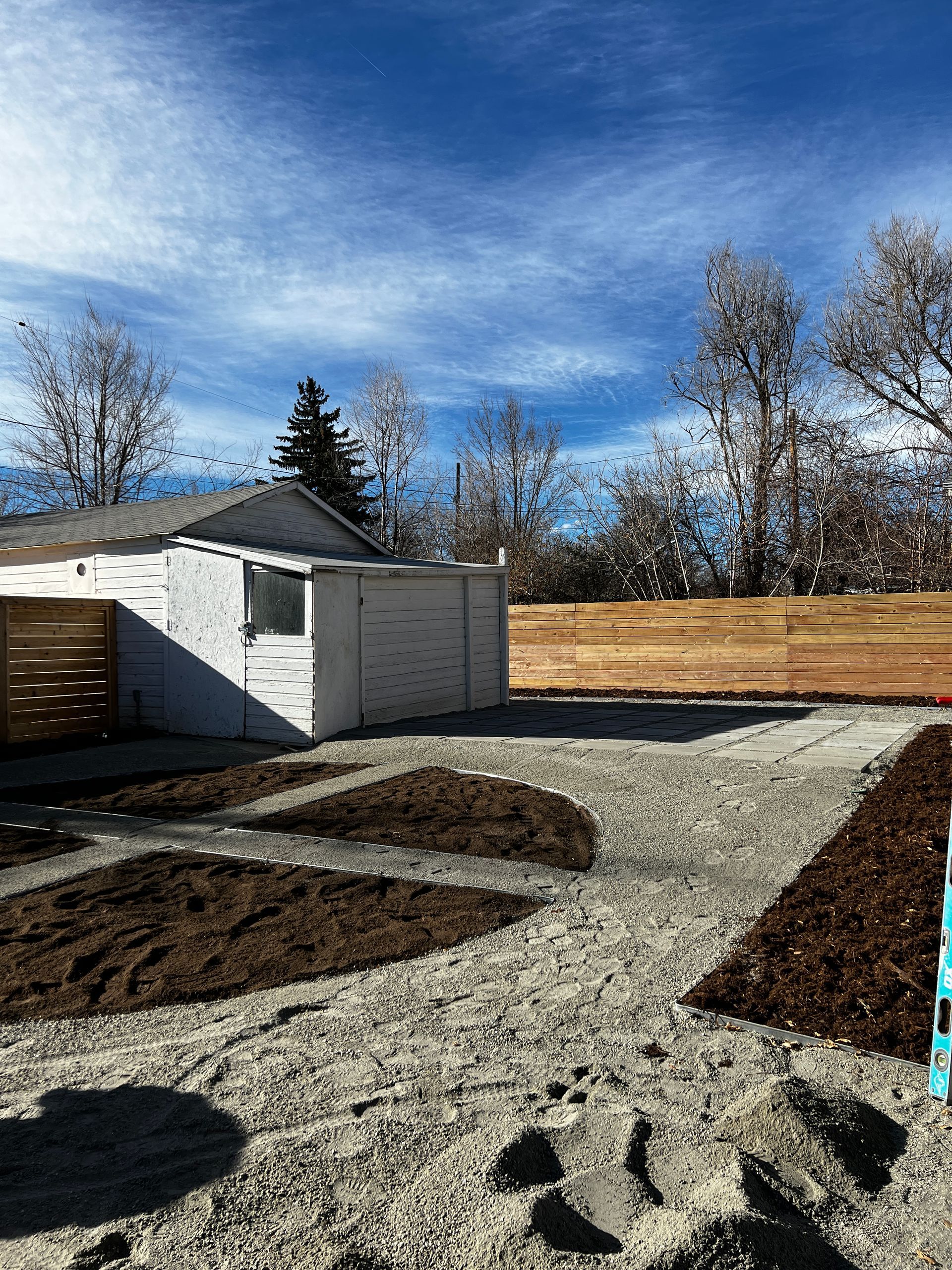 Gravel driveway with mulched planting beds, white shed, and wooden fence against a bright blue sky.