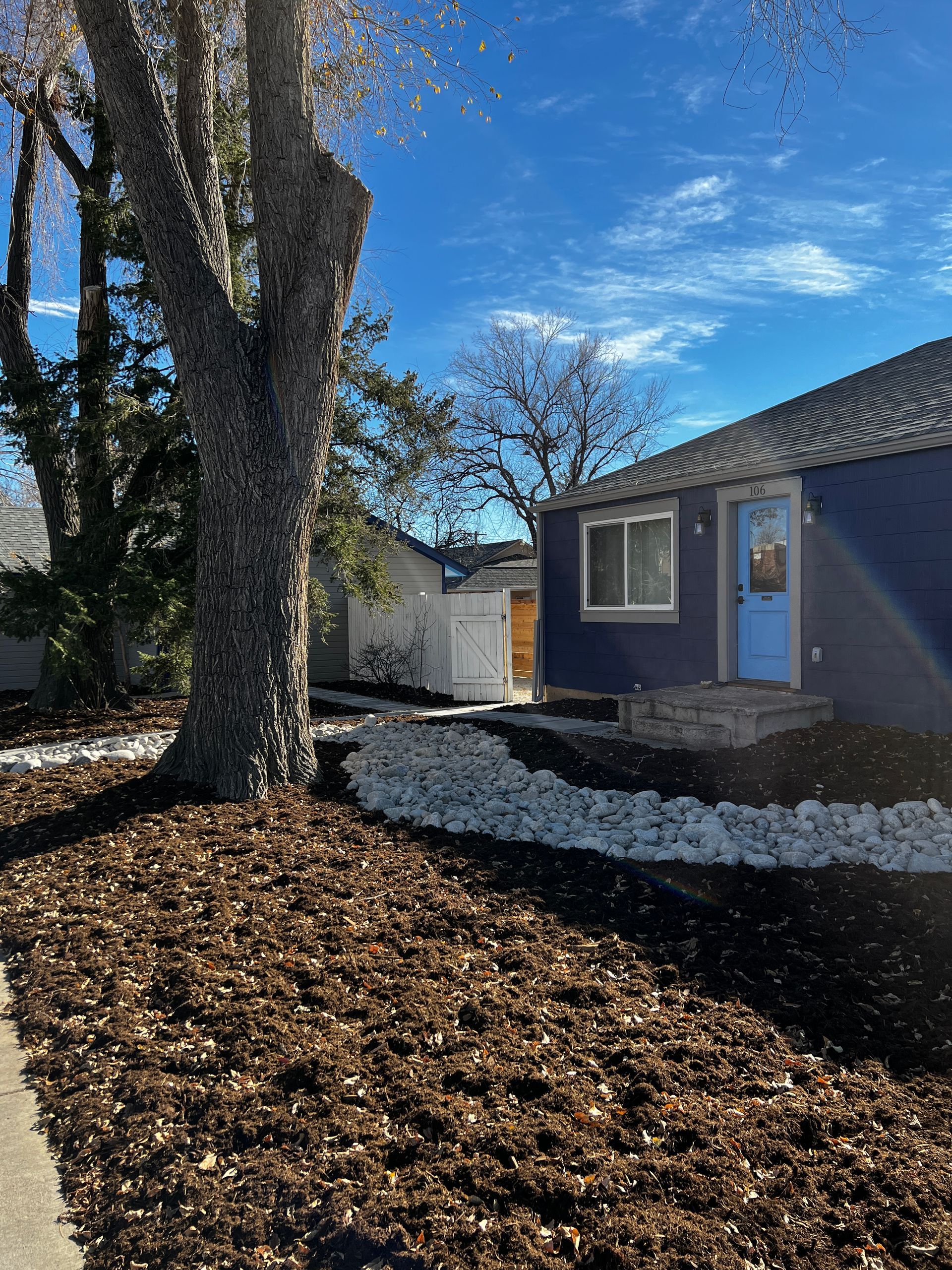 A dark blue house with light blue door and window, brown mulch landscaping, tree, and sunny blue sky.