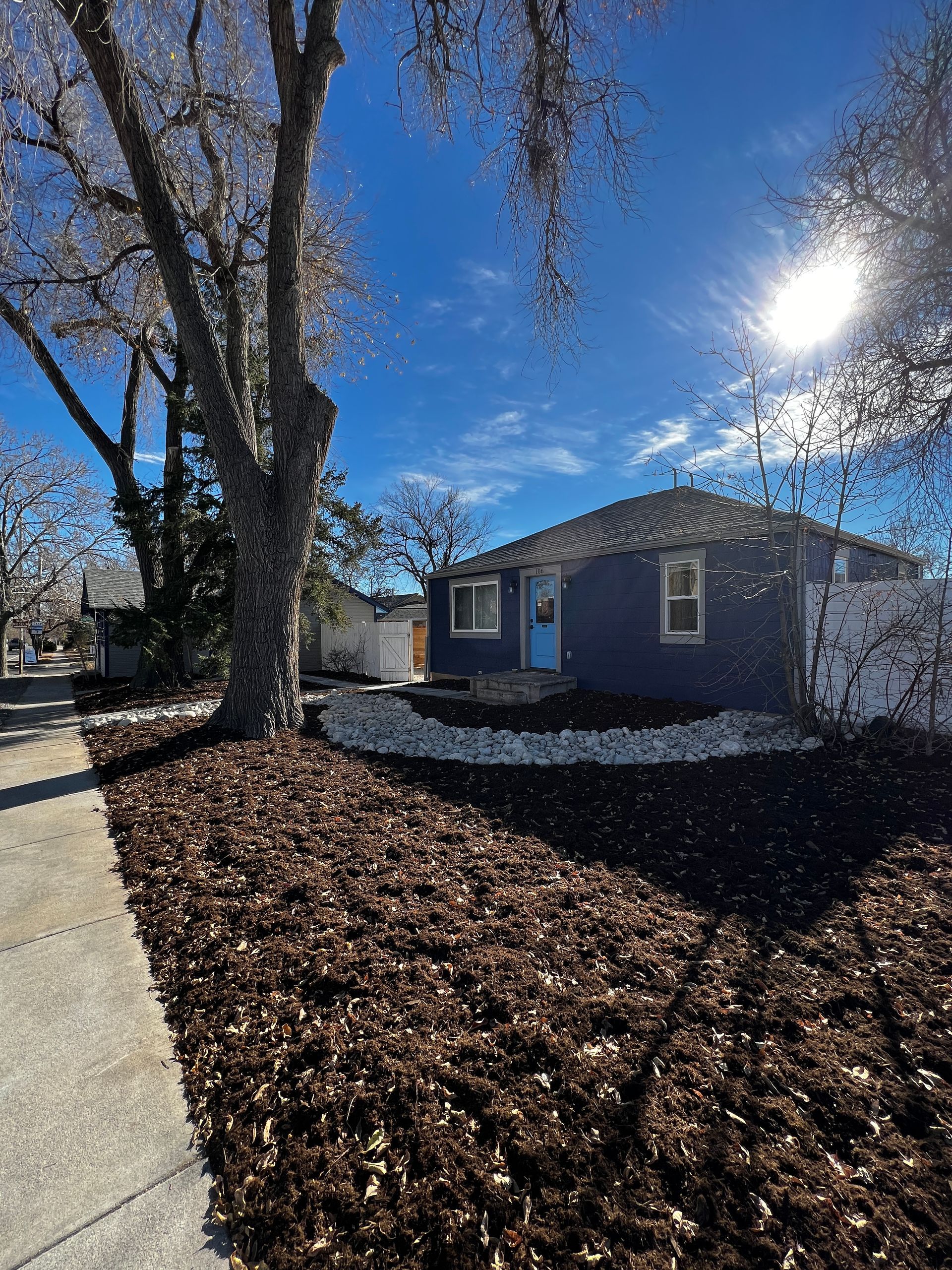 Blue house with a blue door, brown mulch and a tree against a sunny blue sky.