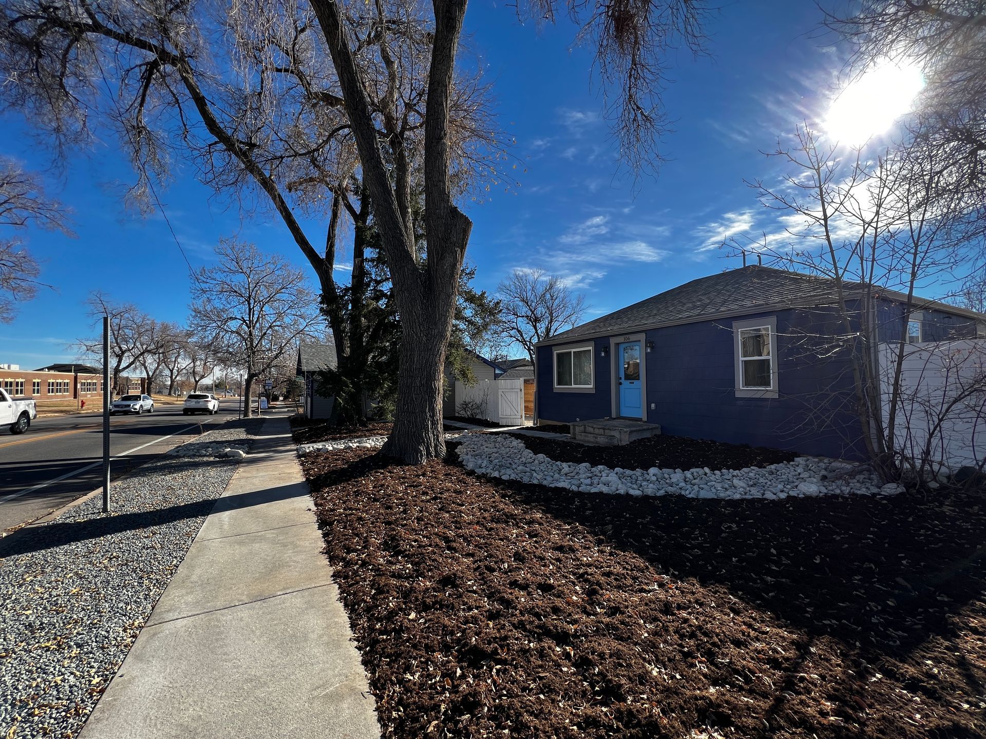 Blue house with a blue door, a sidewalk, trees, and mulch in front. Bright, sunny day.