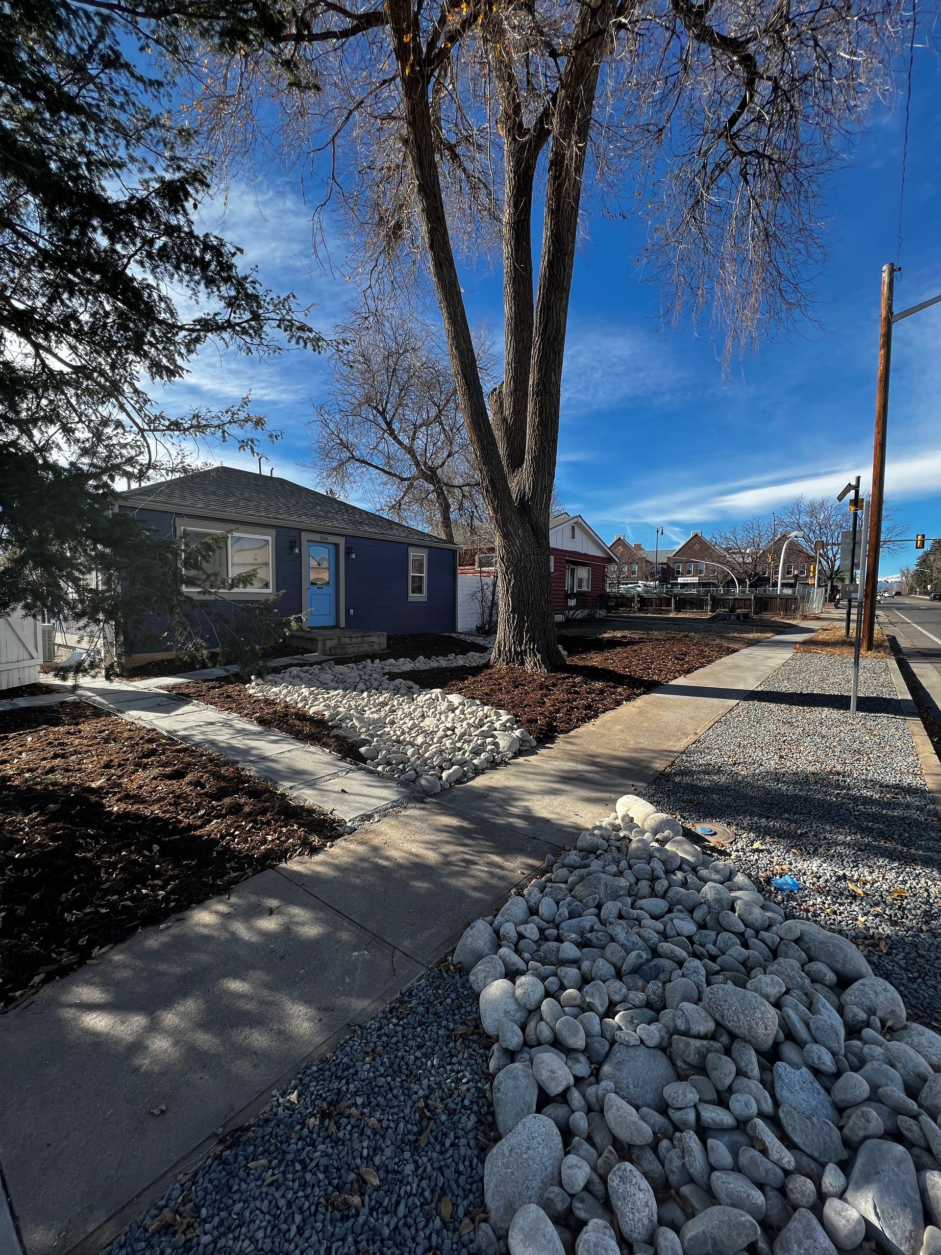Street view: blue house, tree, sidewalk, gravel landscaping. Clear sky.