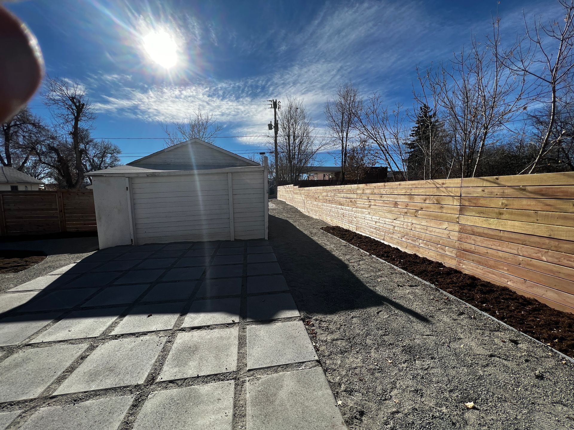 Sunny backyard with a paved patio, gravel pathway, and a wooden fence; a white garage stands in the background.