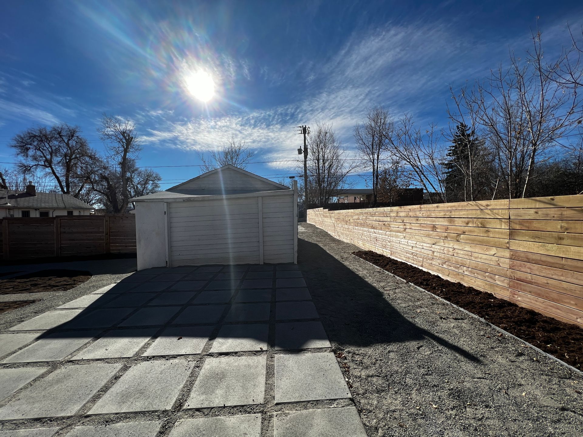 Bright sun over a white garage and stone patio. Gravel path next to a wooden fence under a blue sky.