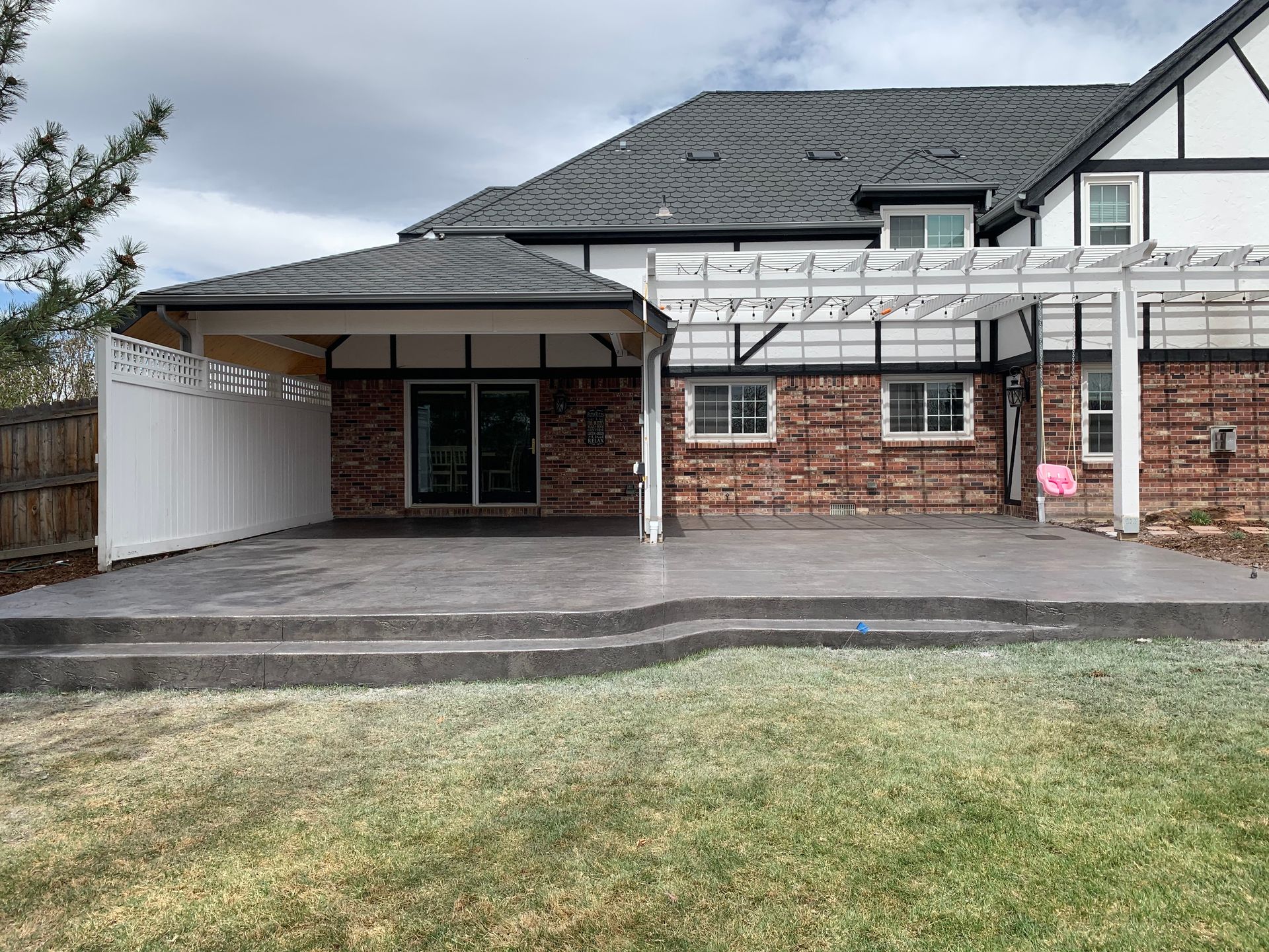 Backyard patio with brick house, pergola, covered seating area, and steps to a lawn.