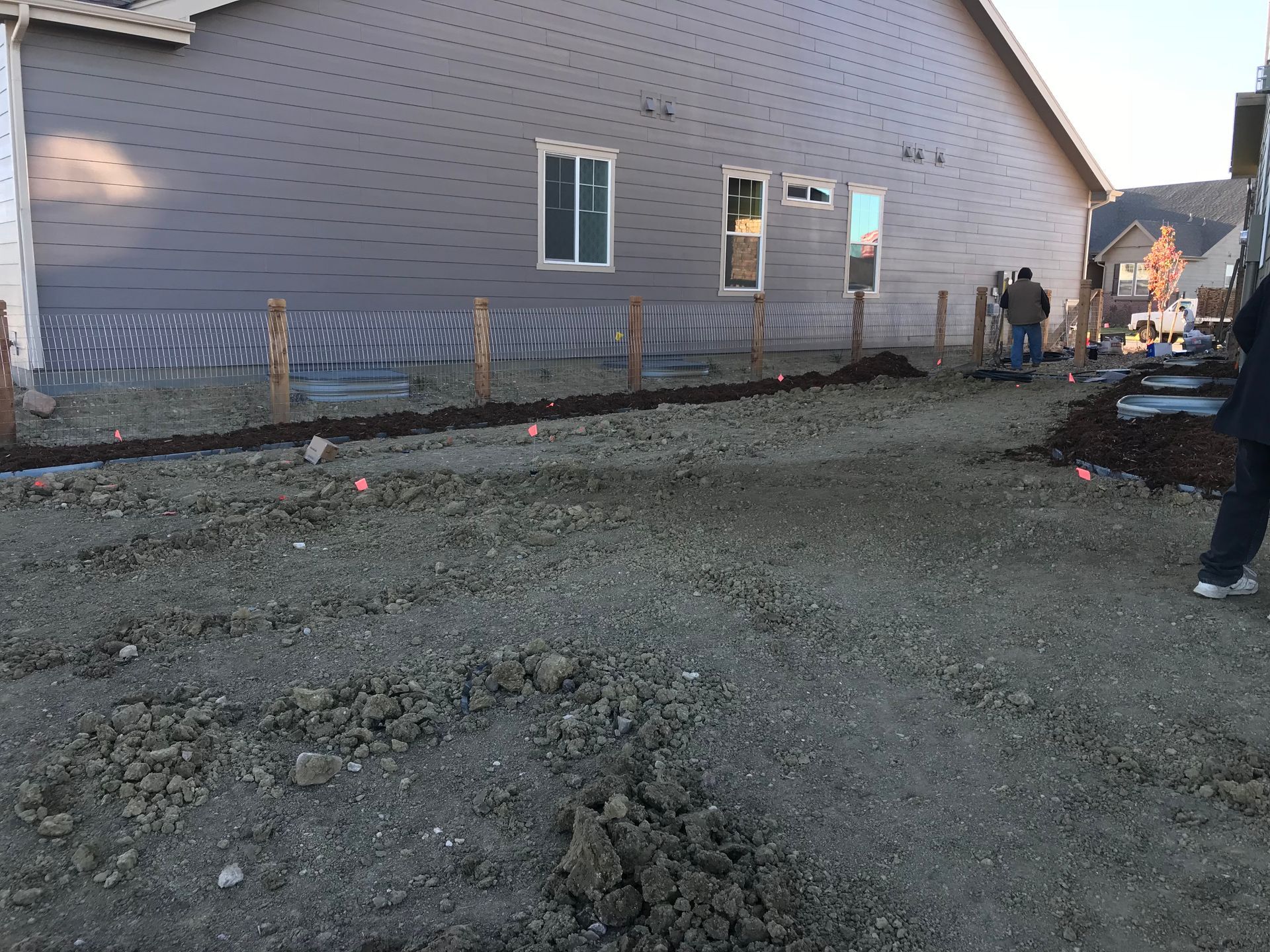 Construction site with gravel ground and a house in the background. Workers are present, and wooden posts mark the area.