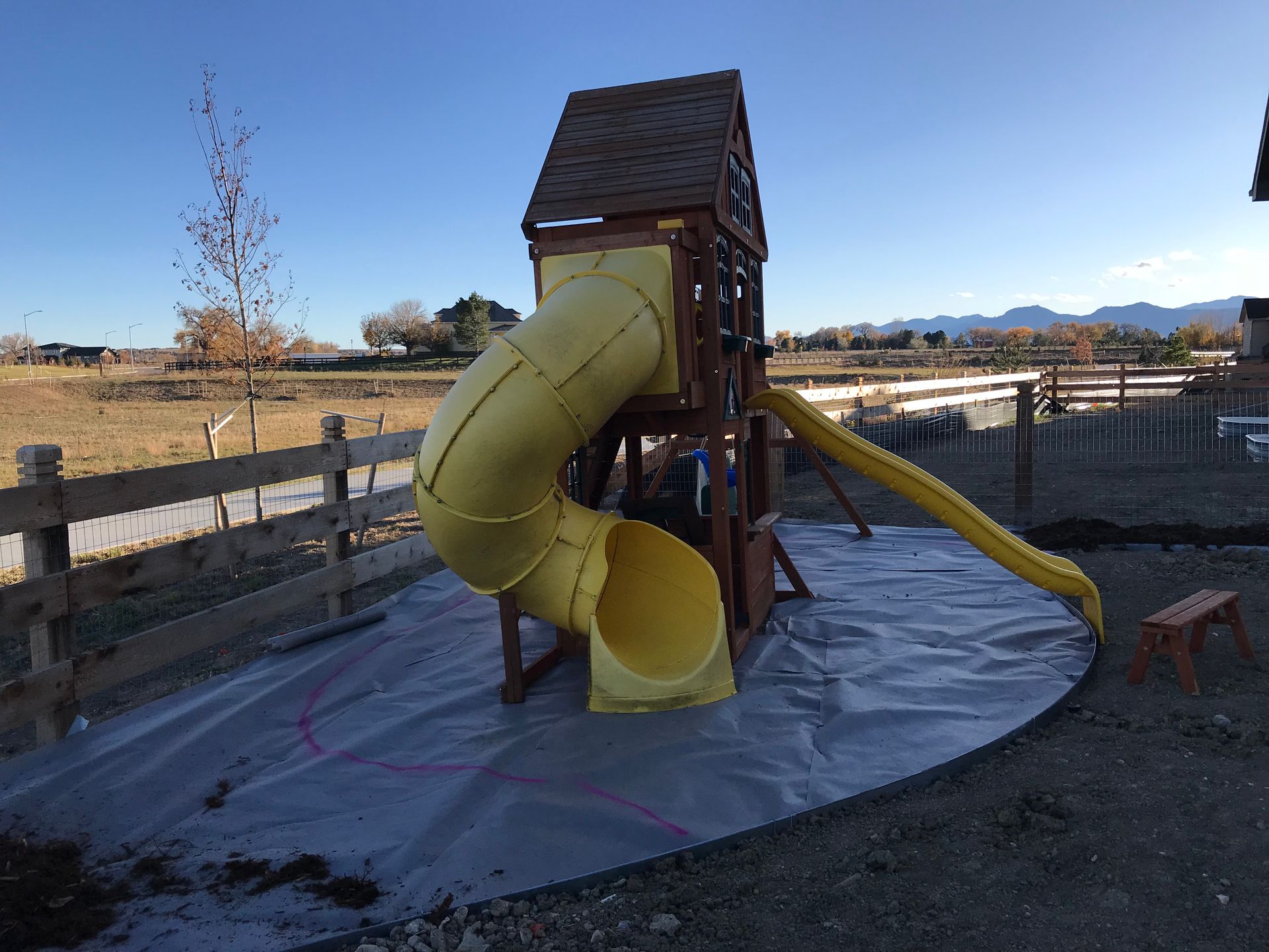 Play set with yellow spiral slide, in a yard with gravel and a fence.