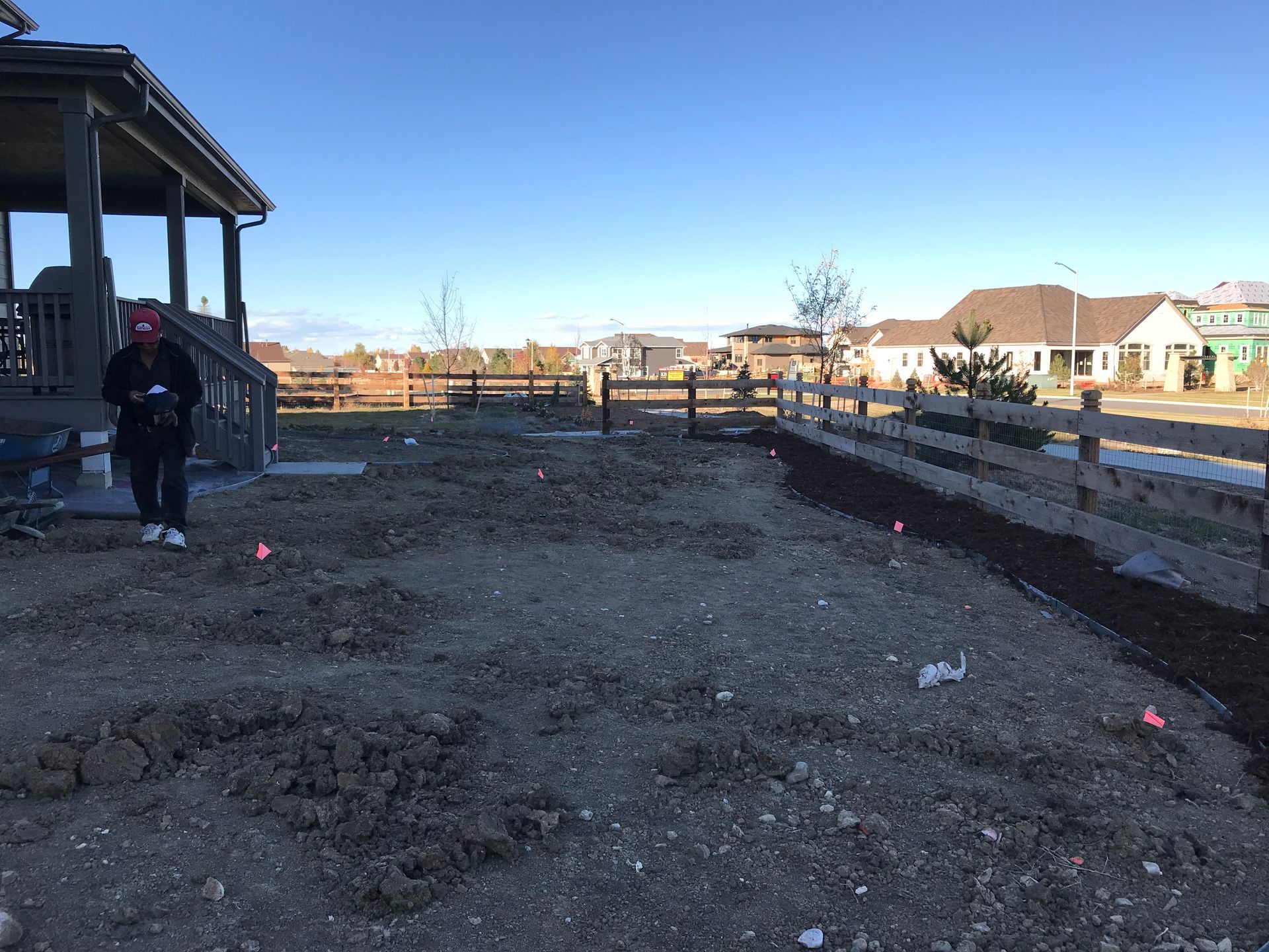 Person standing in a dirt yard with a house on the left, fence, and houses in the background.