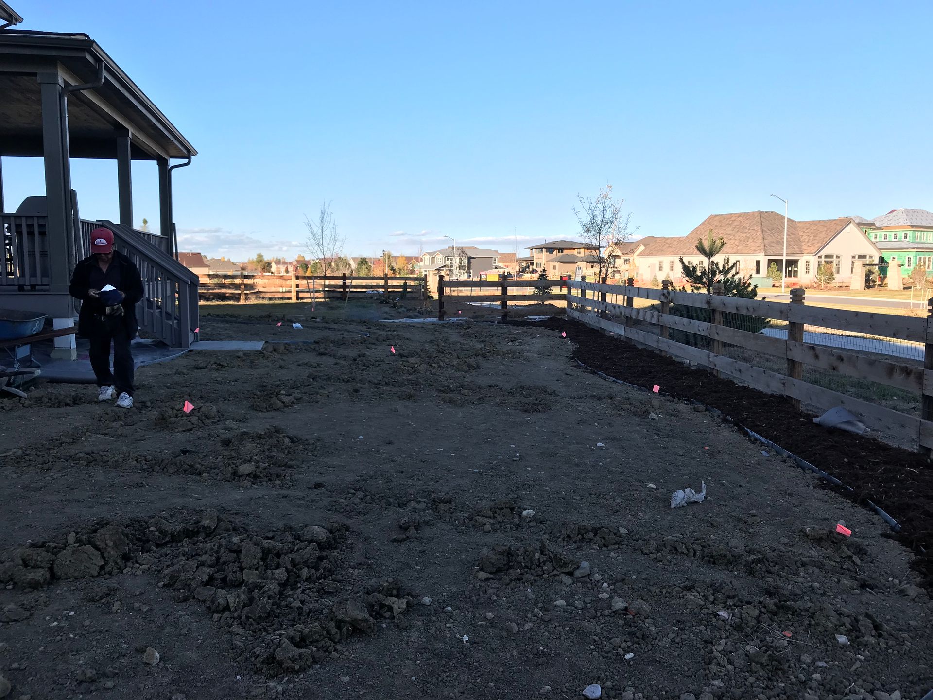 Person surveying a dirt yard with markers. Wooden fence and houses in the background. Sunny day.