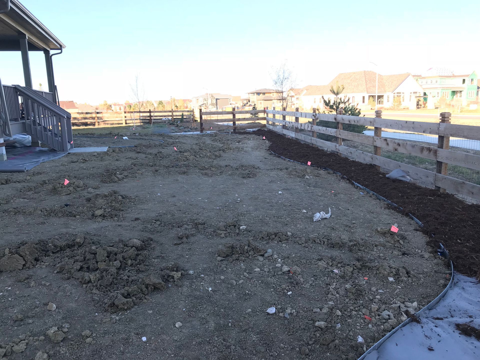 Backyard with fence, dirt ground, small markers, house on the left, and sunny sky.