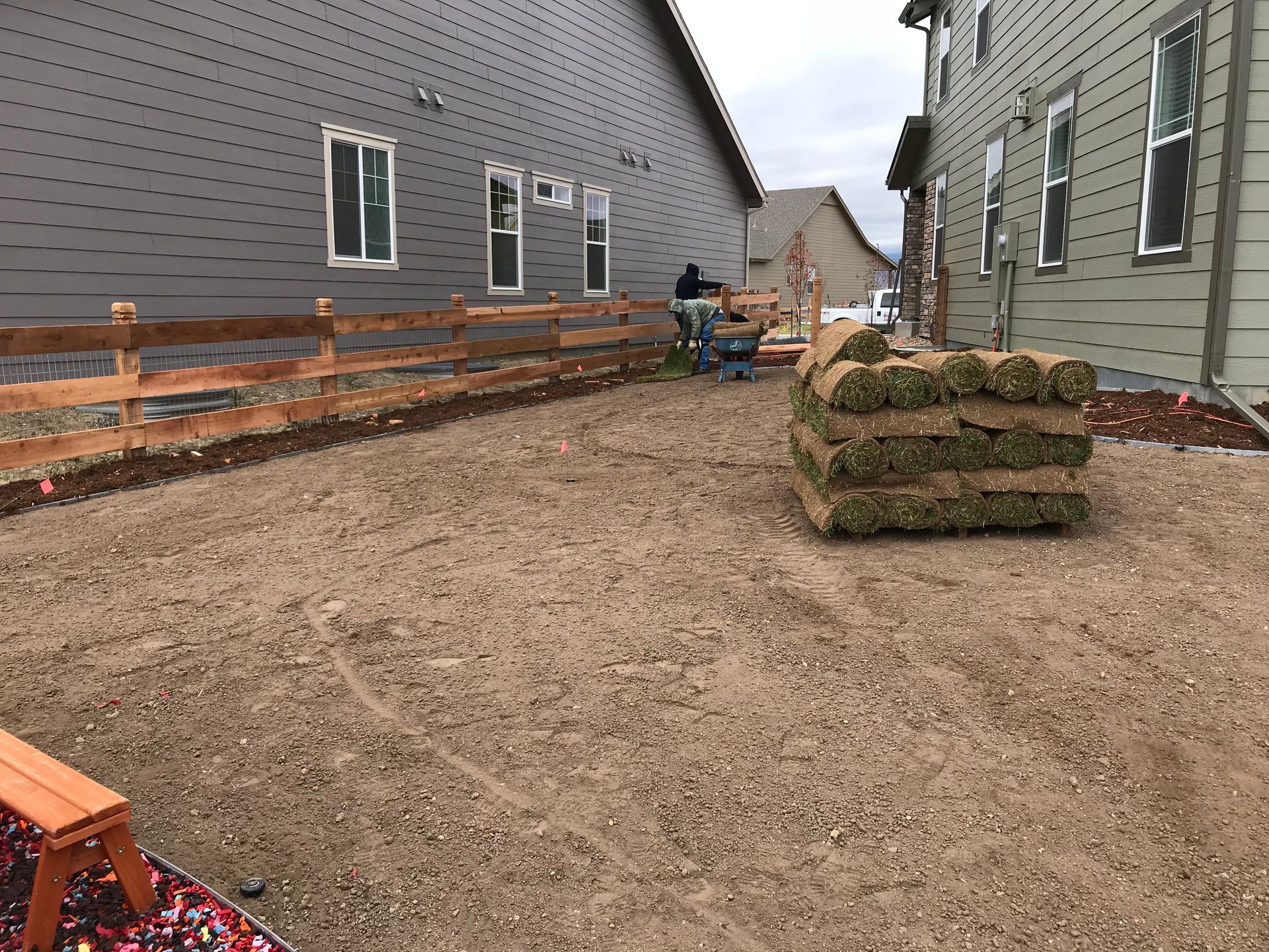 A construction site with rolls of sod, gravel ground, a fence, and workers in the background.