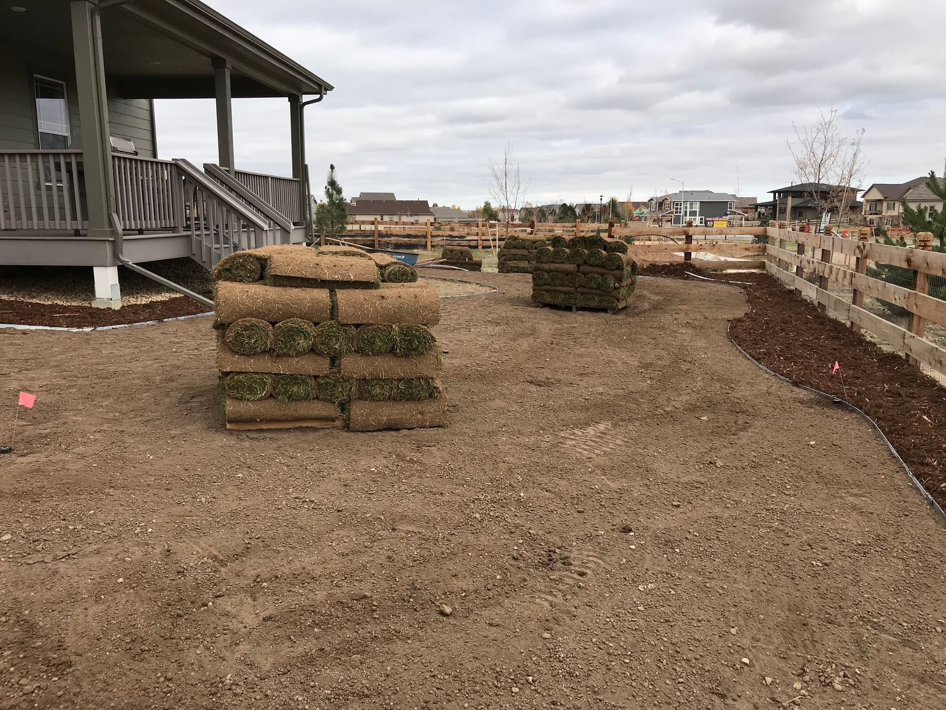 Stacks of sod in a dirt yard near a house with a porch and a wooden fence on a cloudy day.