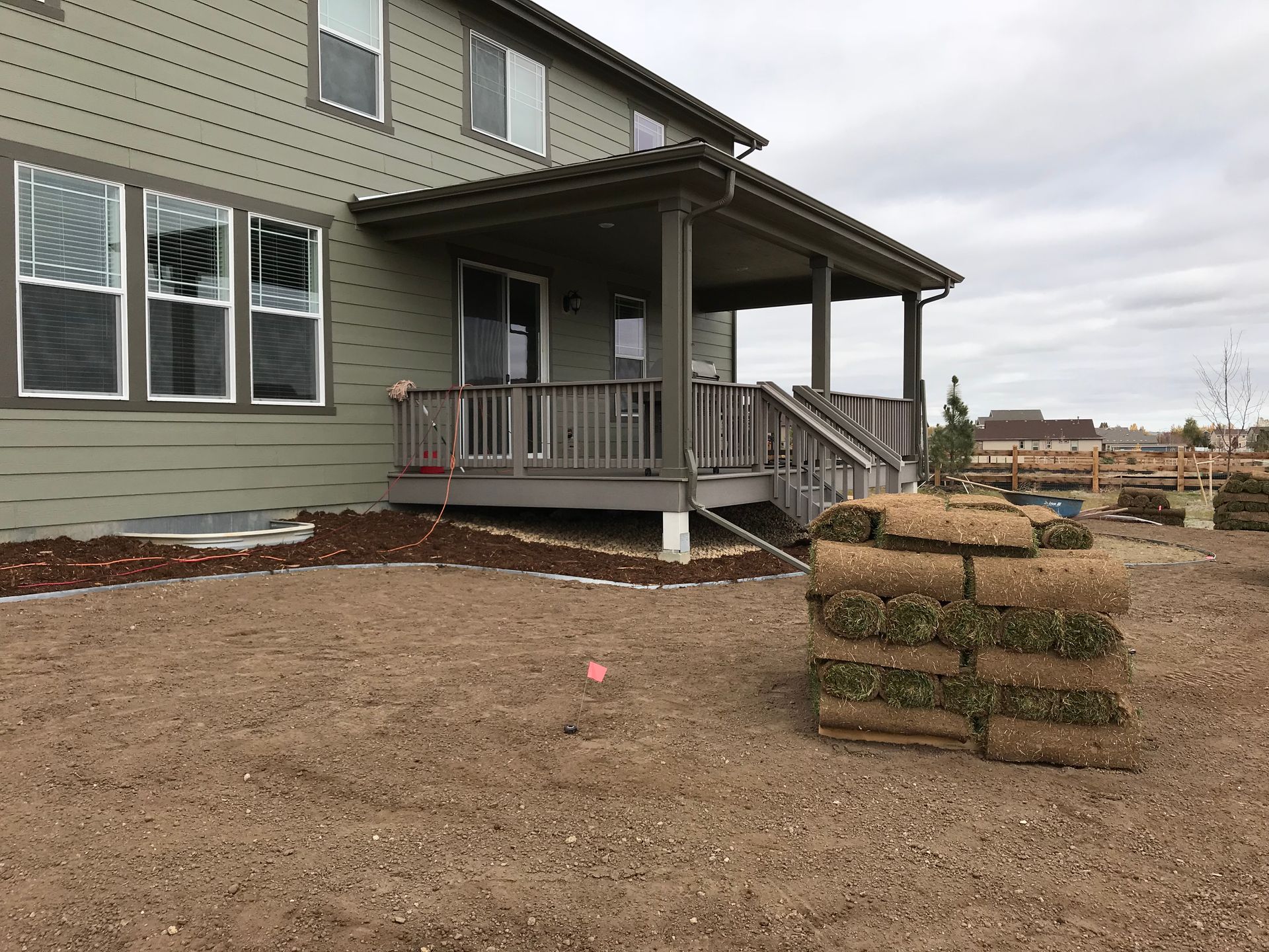 Back of a house with a porch. Stack of sod on the ground. Soil with red marker. Cloudy sky.