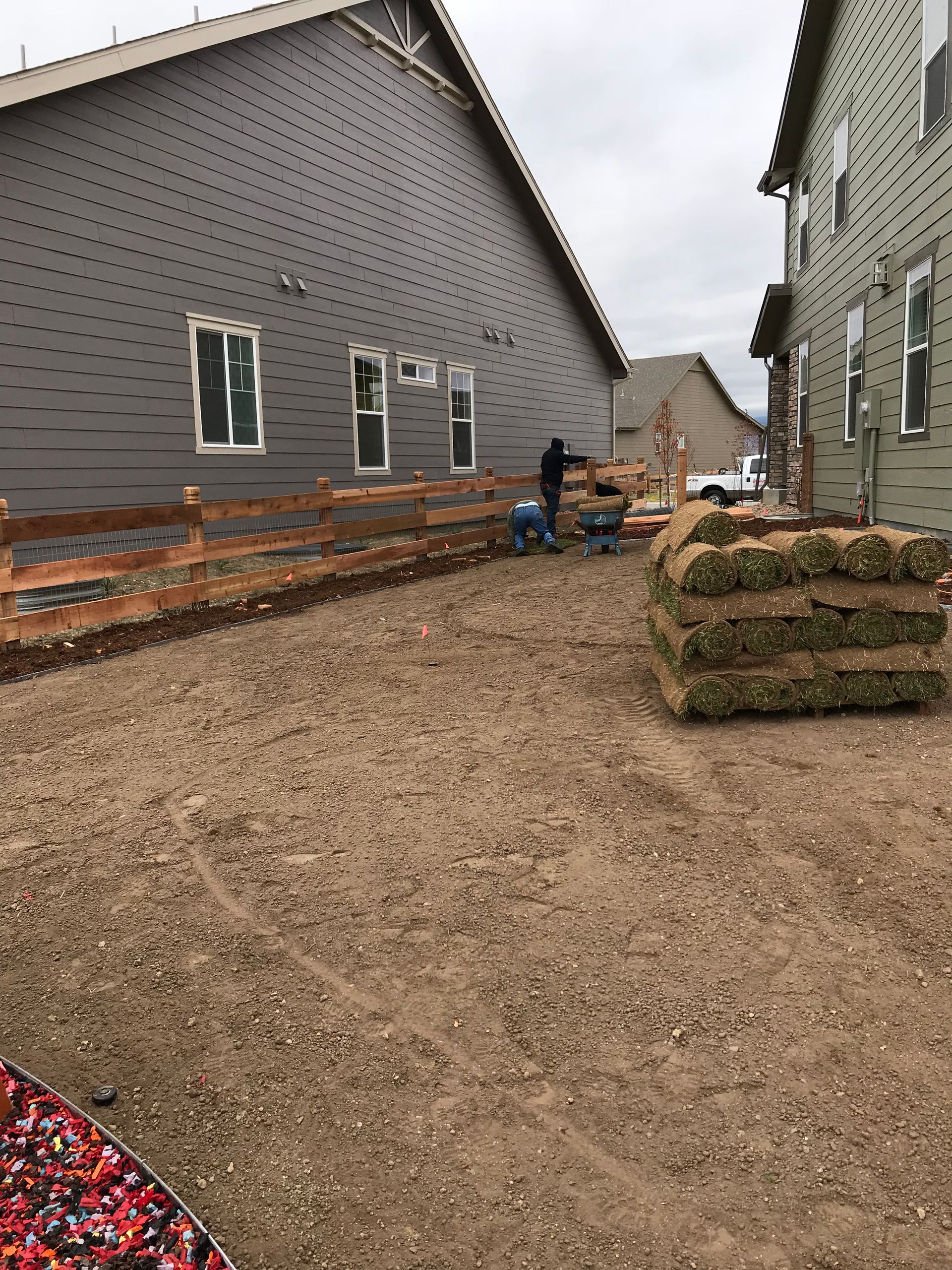 Construction site with dirt, wooden fence, grass rolls, and workers. Gray-sided houses and cloudy sky.