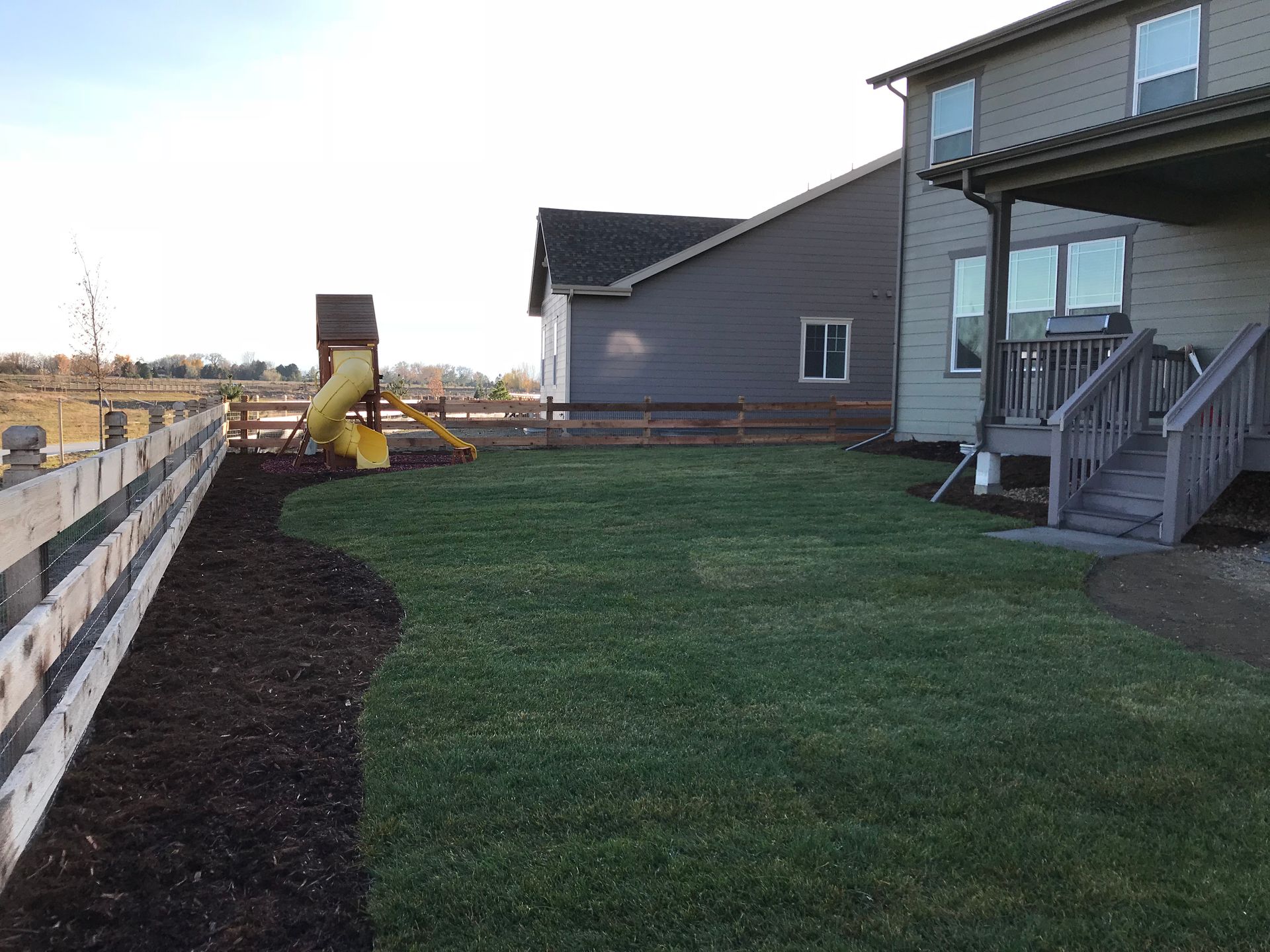 Backyard with a wooden fence, green lawn, and a play structure. A house is on the right.