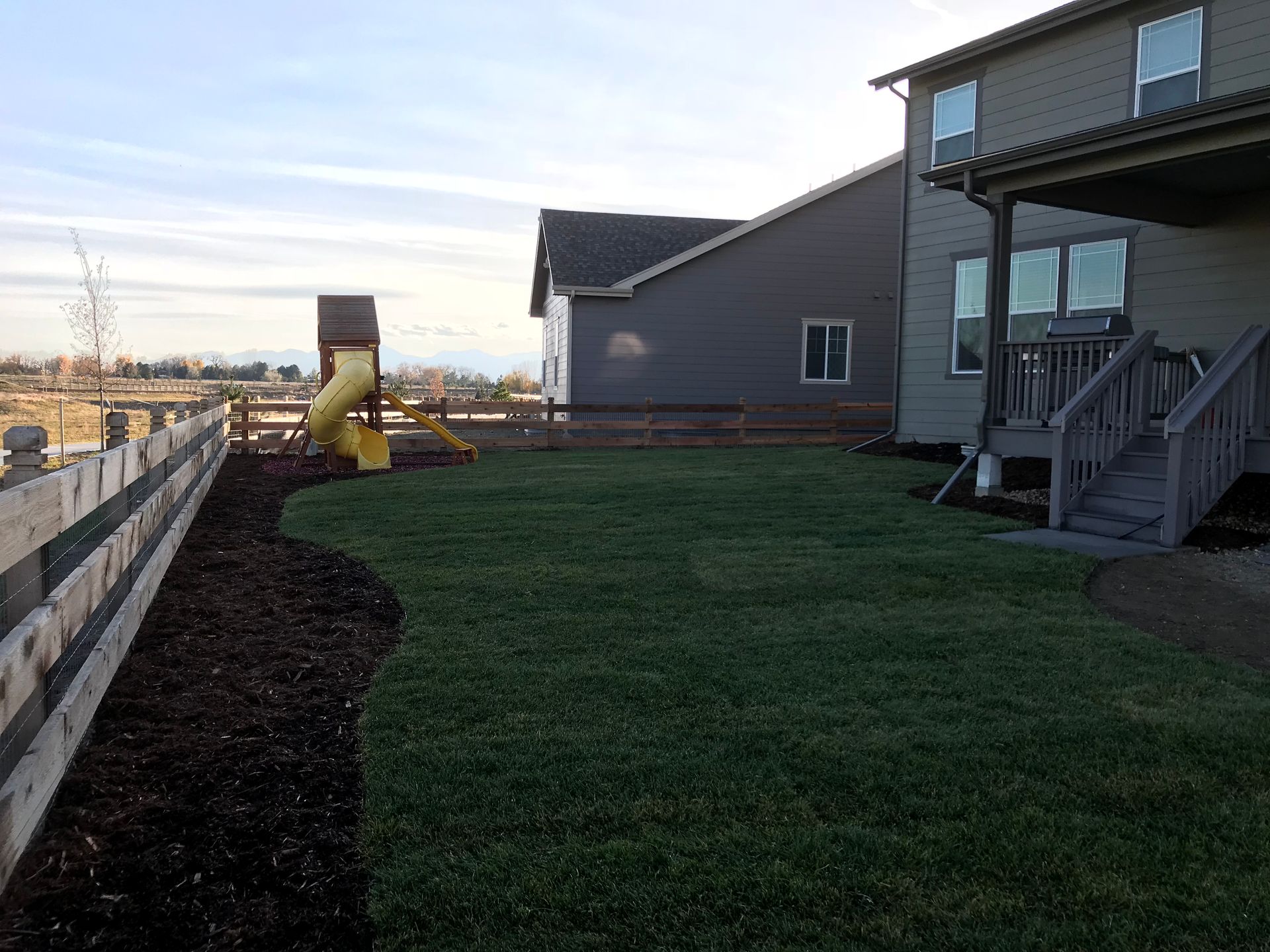 Backyard with lush green grass, a playground, and a wooden fence, next to a two-story house.