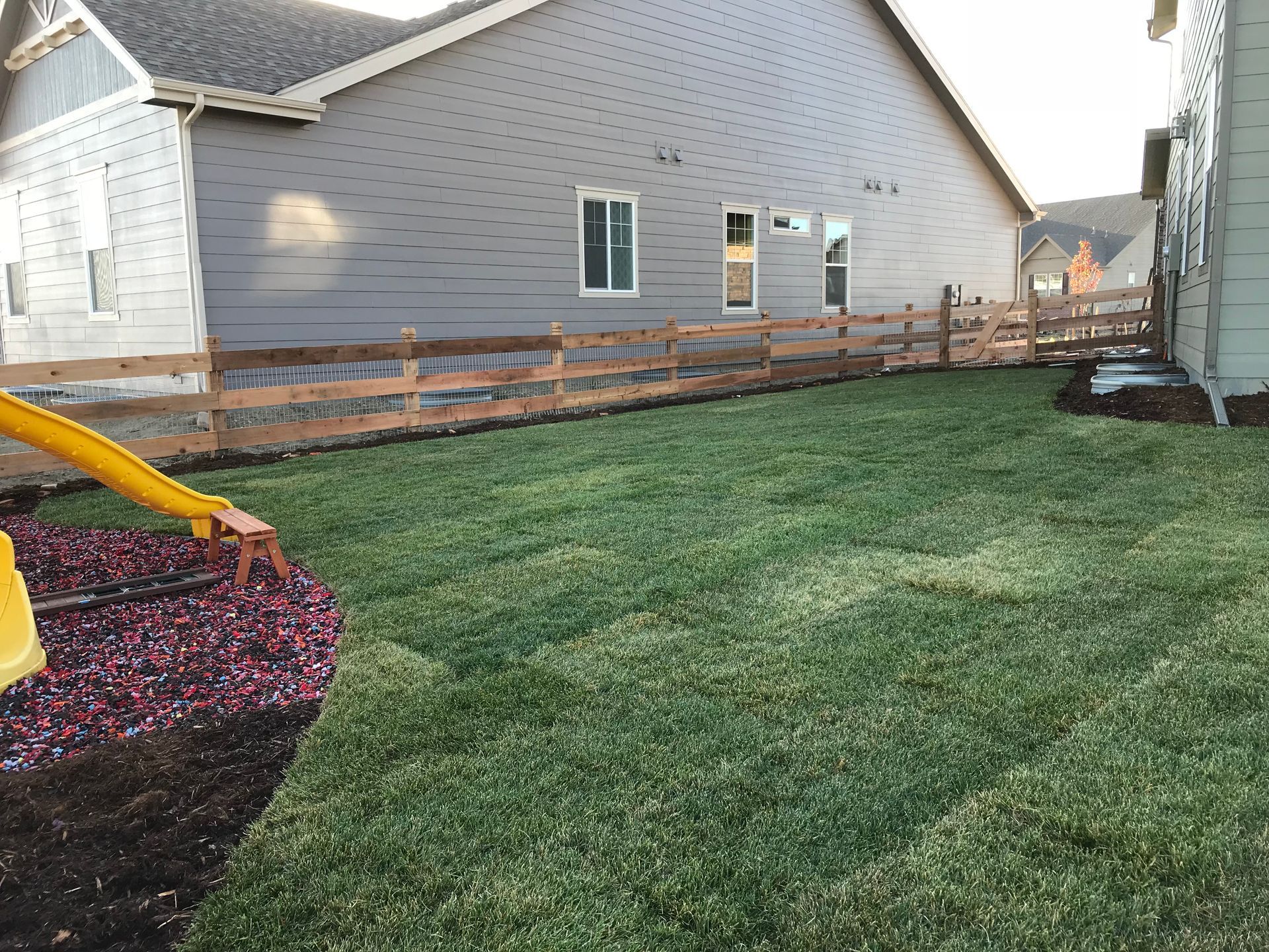 A grassy backyard with a wooden fence. A house is in the background. A playground is to the left.