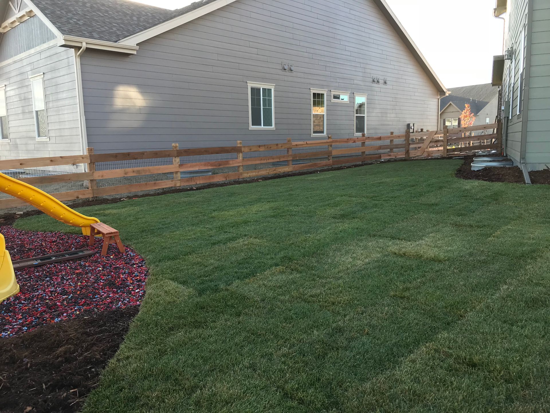Backyard with green lawn, wooden fence, gray houses, and mulch bed.