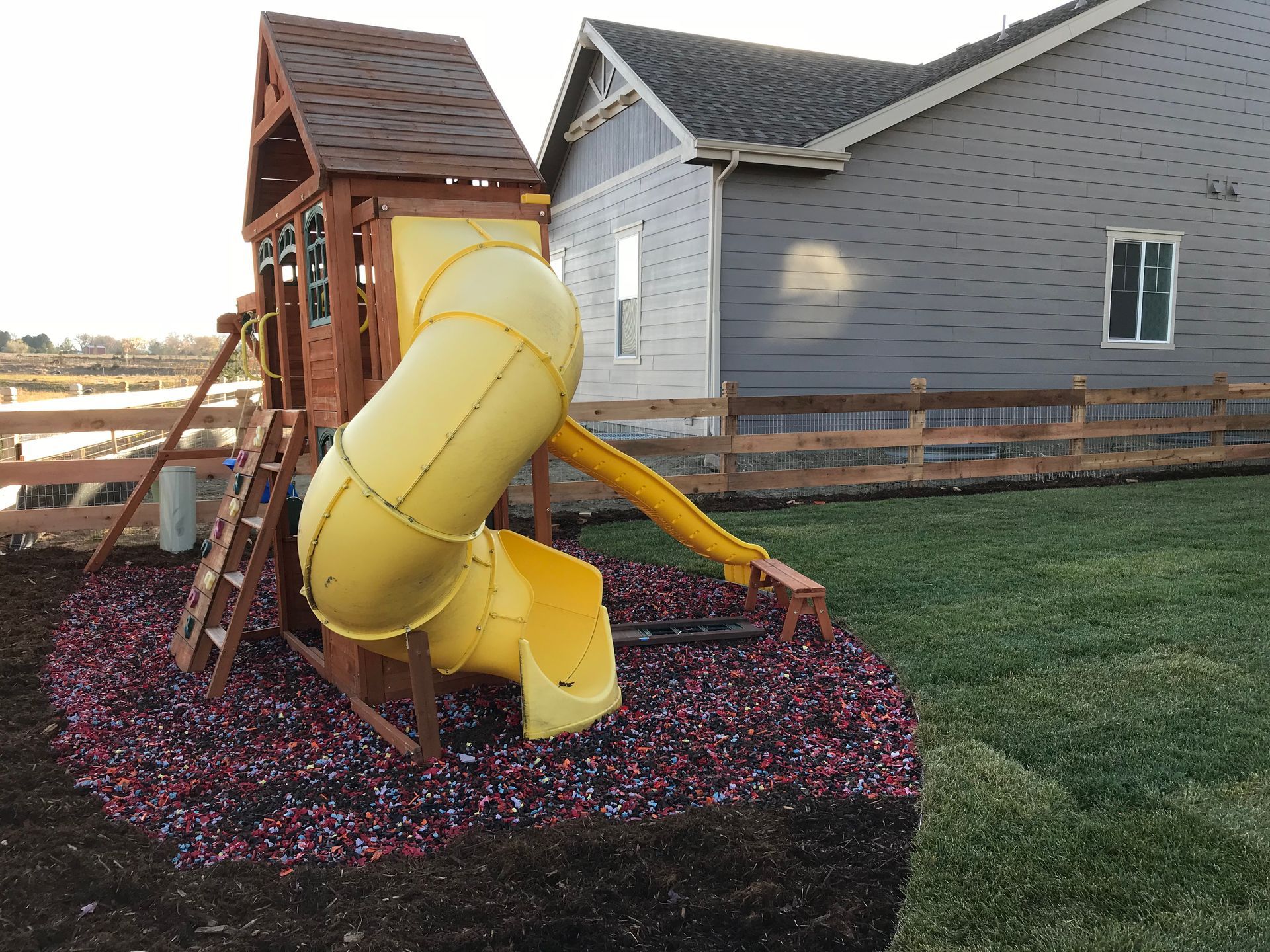Playground with yellow slide, brown playhouse, and gray house in backyard.
