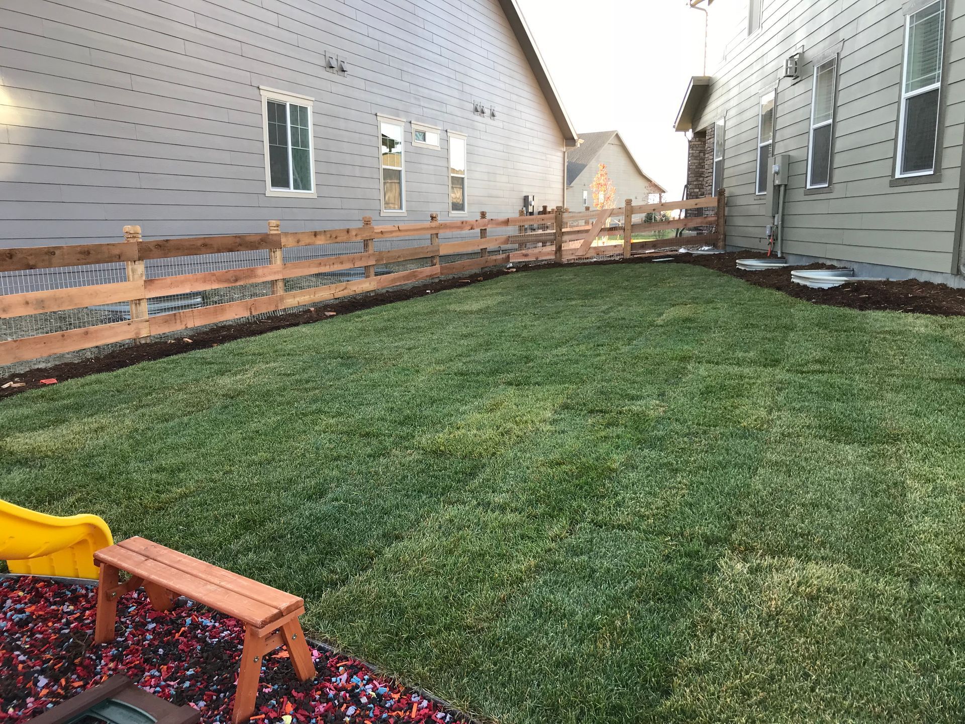 Lawn between two houses, wooden fence, small wooden bench, and a yellow slide.