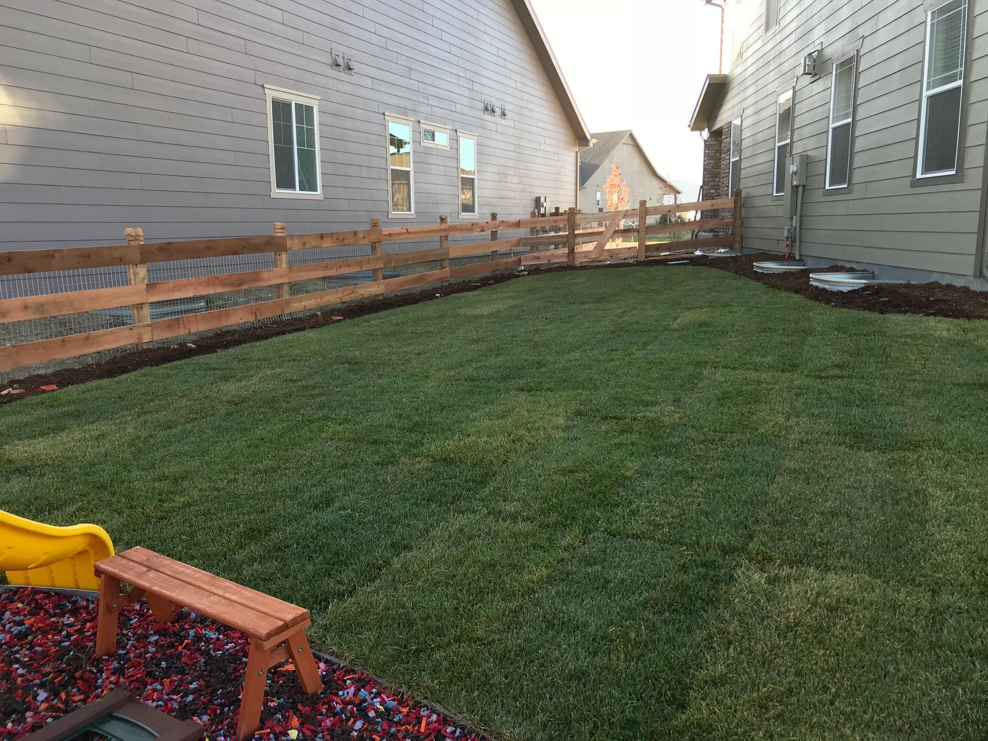 Lawn bordered by a wooden fence between two grey houses; a small playground bench is in the lower left corner.