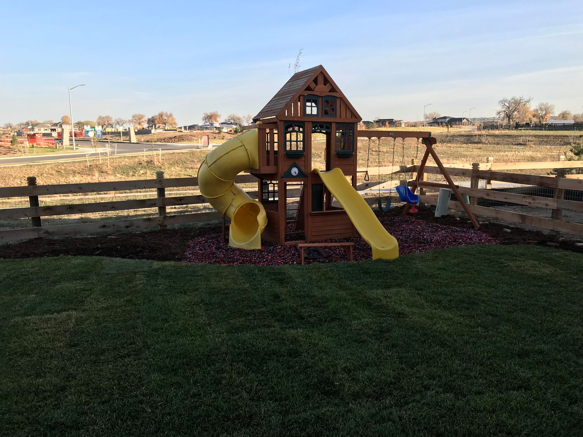 Wooden playground with yellow slide and swing set in a grassy yard, under a blue sky.
