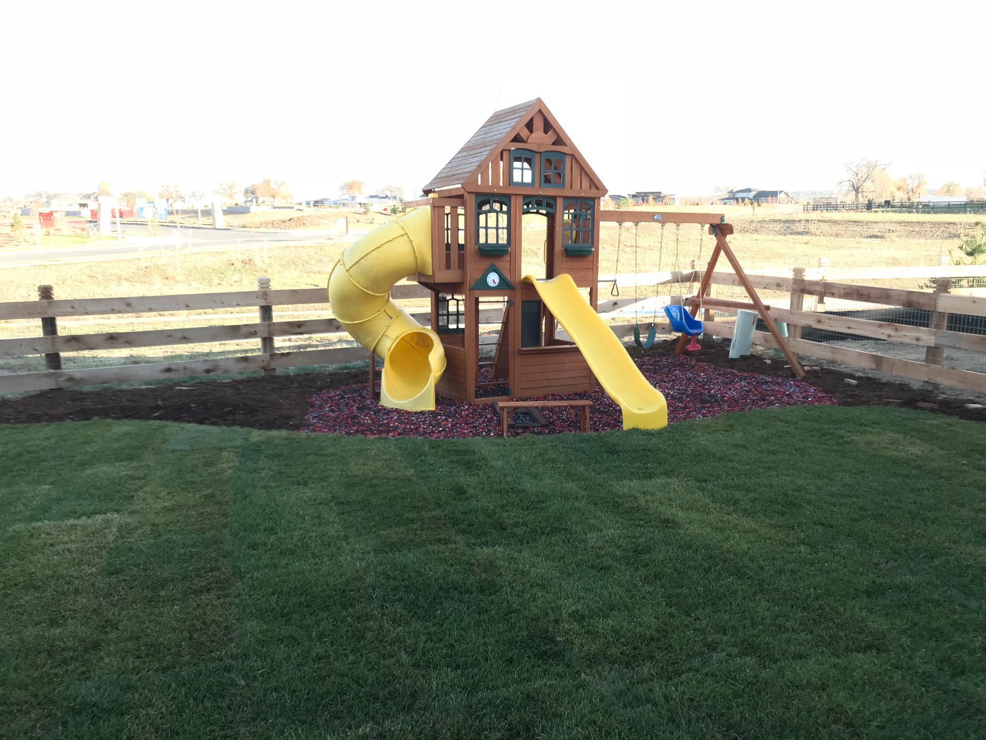 Playground set with yellow slide and tunnel on mulch, surrounded by grass and a fence.