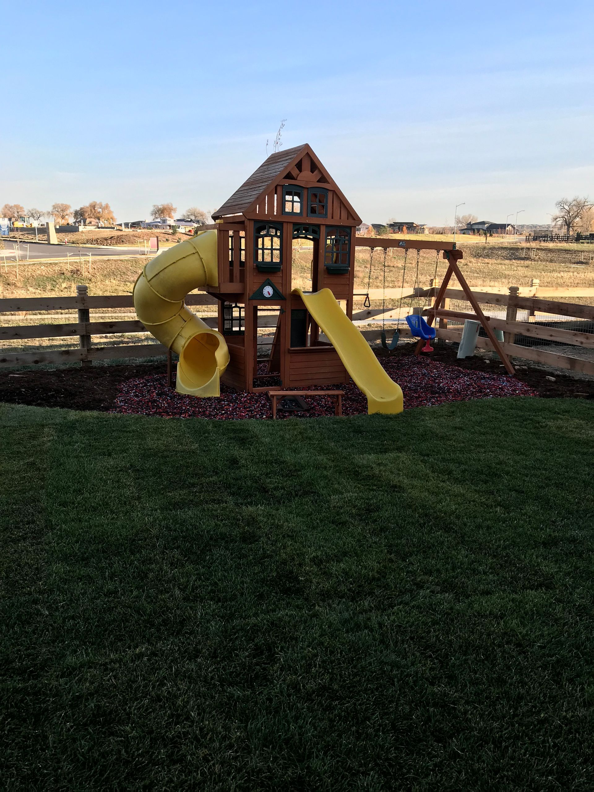 Wooden playset with yellow slide and tunnel, surrounded by wood fence, grass, and a brown field.