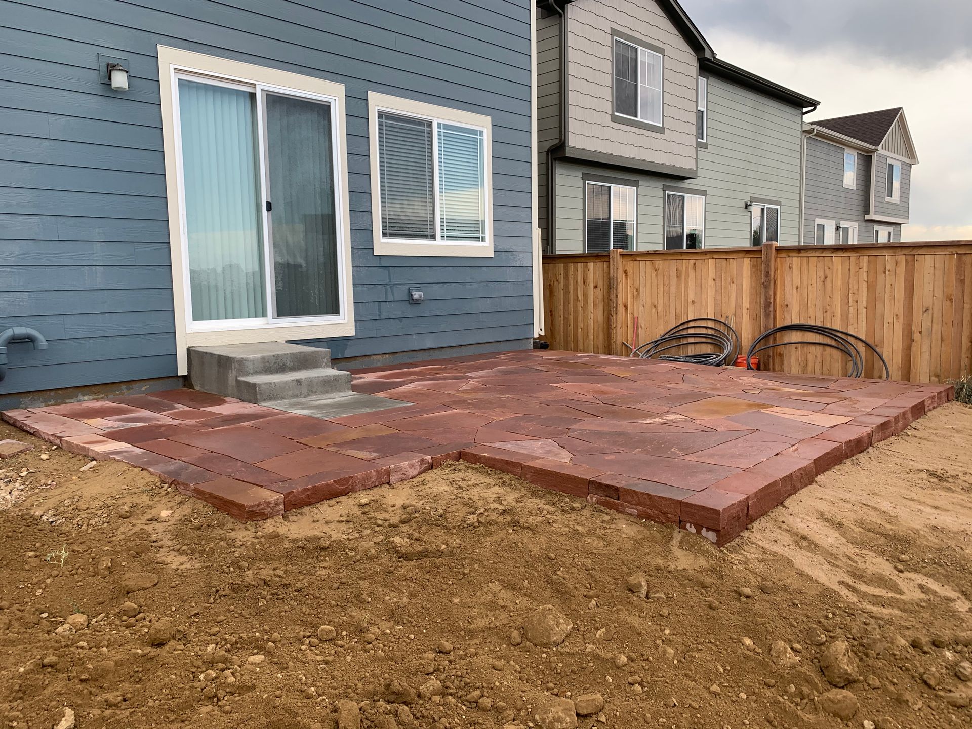 A newly built red paver patio outside a blue house with a sliding door and a wooden fence.
