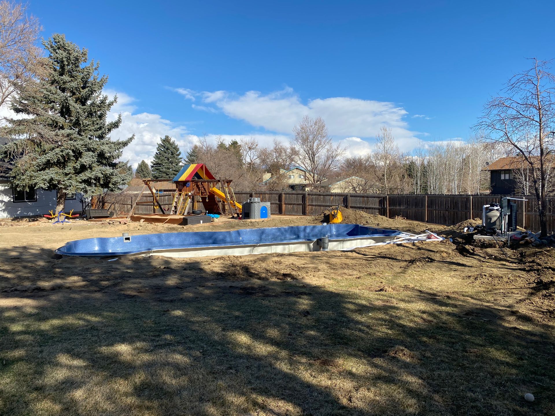Backyard with an unfinished pool, play structure, and excavator under a blue sky.