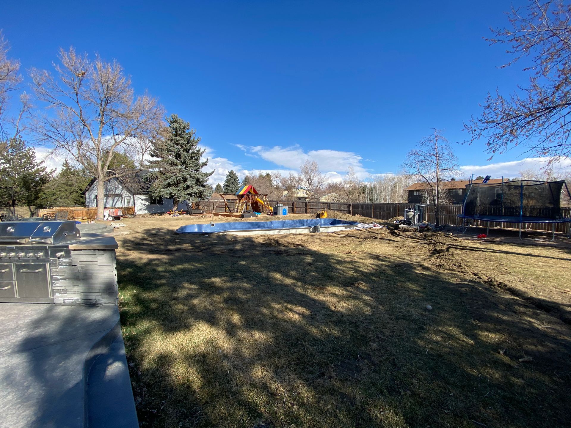 Backyard with grass, pool cover, grill, trampoline, and play structure under a blue sky.