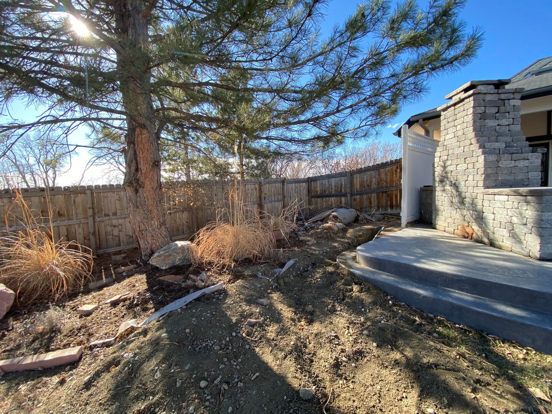 Backyard with wooden fence, tree, rocks, and a portion of a house with stone accents.