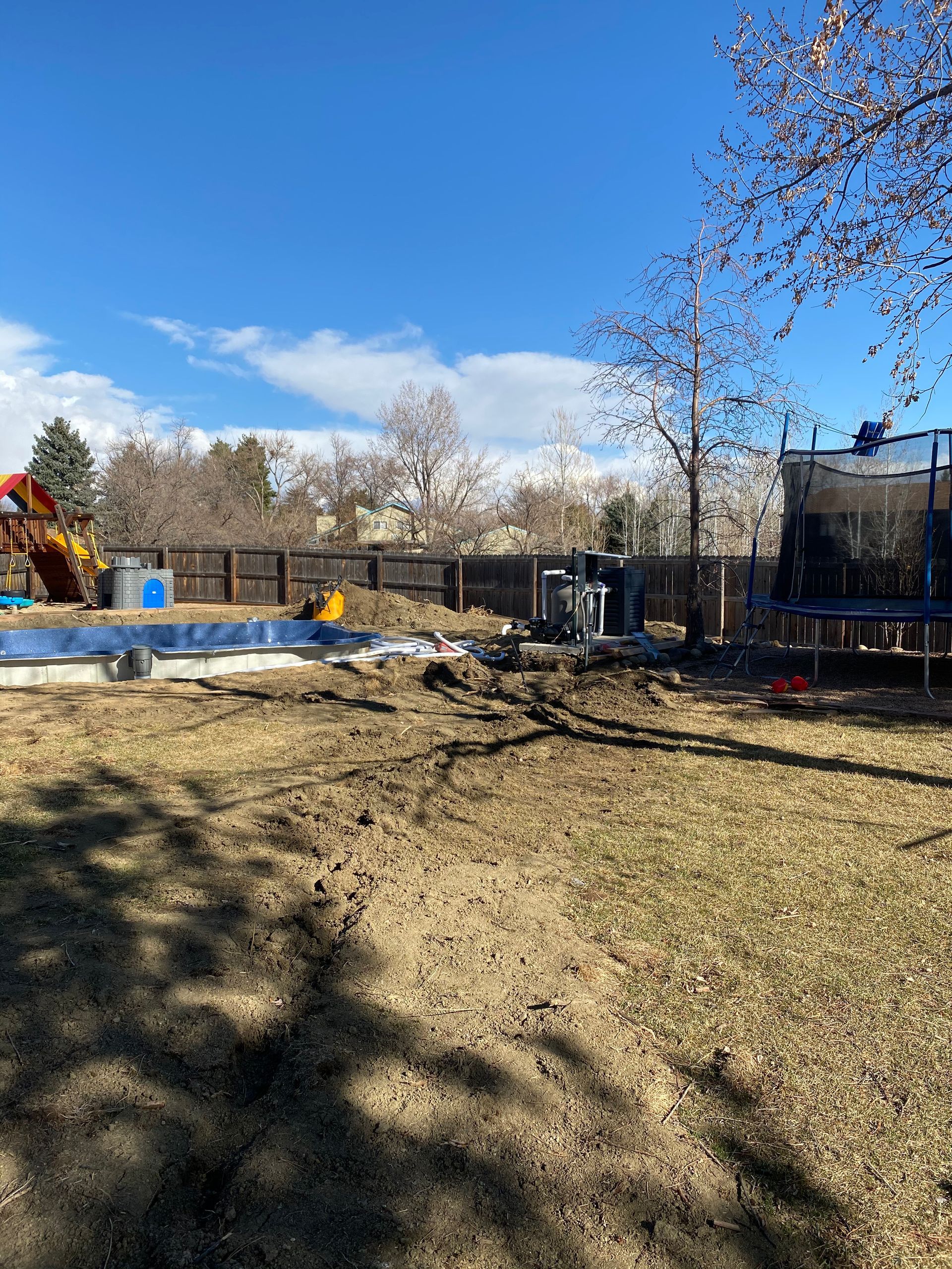 Backyard with a pool under construction, a trampoline, and a playground under a blue sky.
