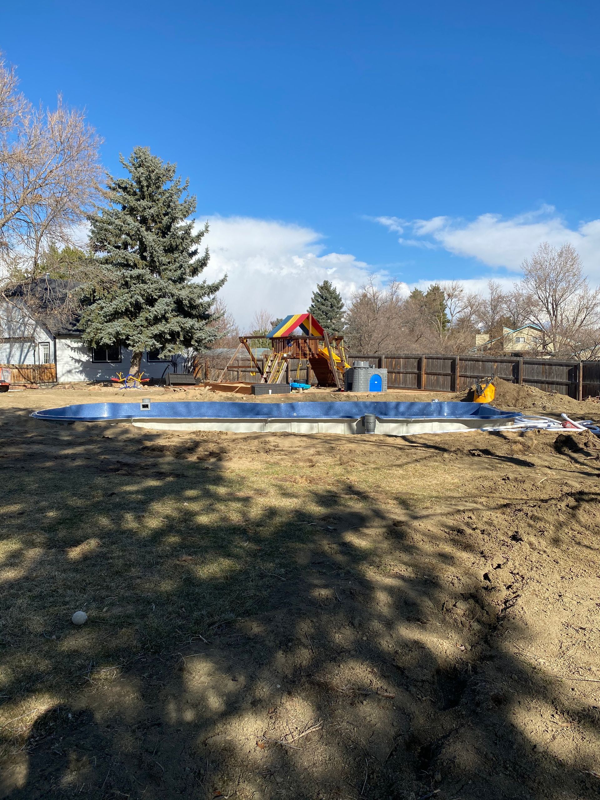 Backyard with a blue concrete pool frame, a wooden play structure, and sparse brown grass under a blue sky.