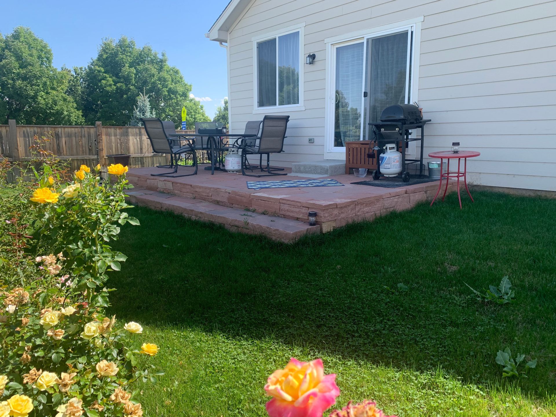Backyard patio with table and chairs, grill, and roses. Green grass and blue sky.