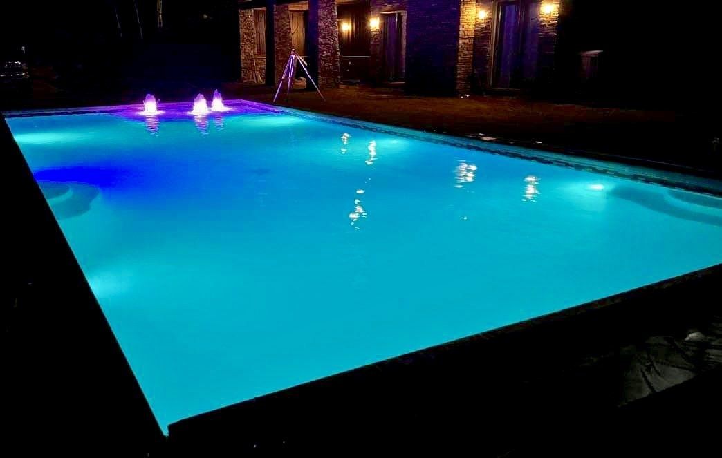 A rectangular pool illuminated with blue lights at night. Fountains and a stone structure are in the background.