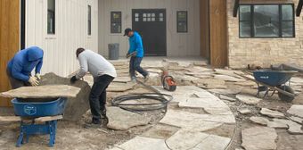 Construction workers laying stone pavers on a patio outside a house.