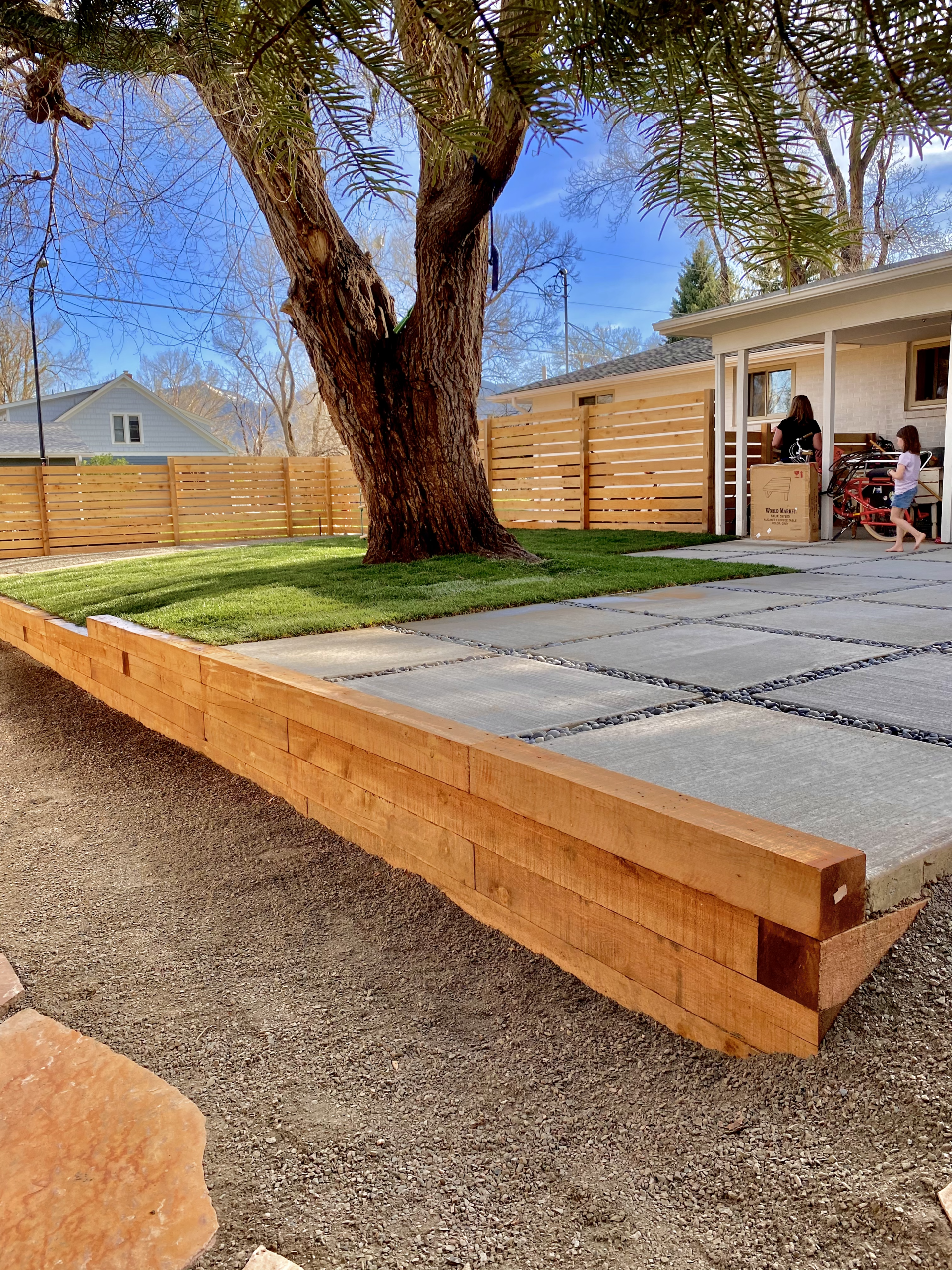 Wooden retaining wall, patio, and fence in a yard with a large tree. Children are playing near the house.