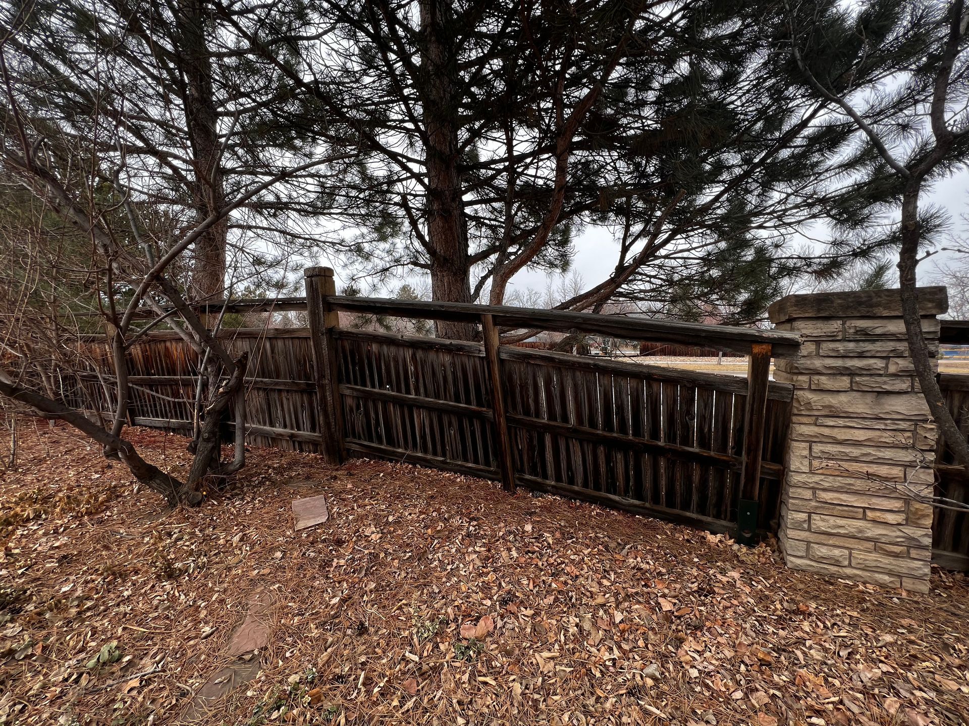 Rustic wooden fence with brick pillar, surrounded by trees and fallen leaves.