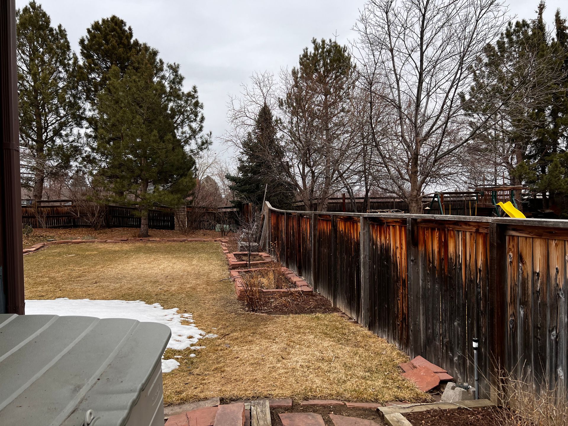 Backyard scene with brown fence, trees, patches of snow, and a playground visible.