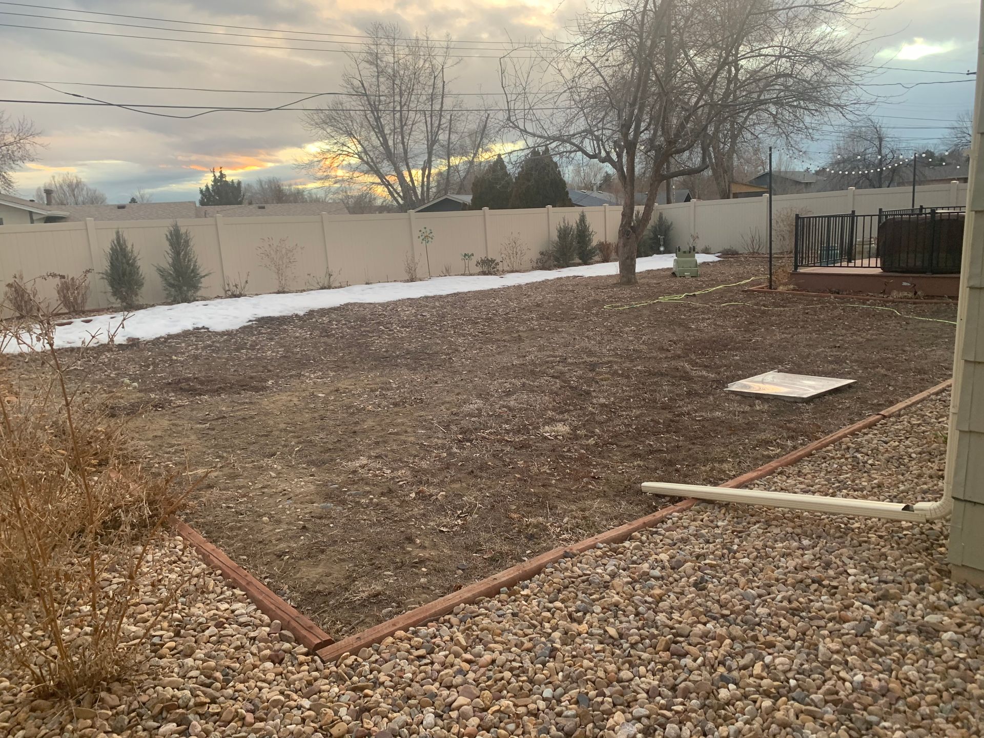 A backyard with snow, brown gravel, a wooden deck, and a fence under a cloudy sky.