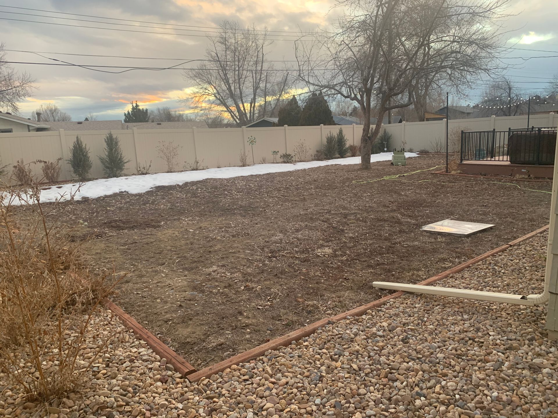 Backyard with brown dirt, snow, tan fence, and bare trees under a cloudy sky.