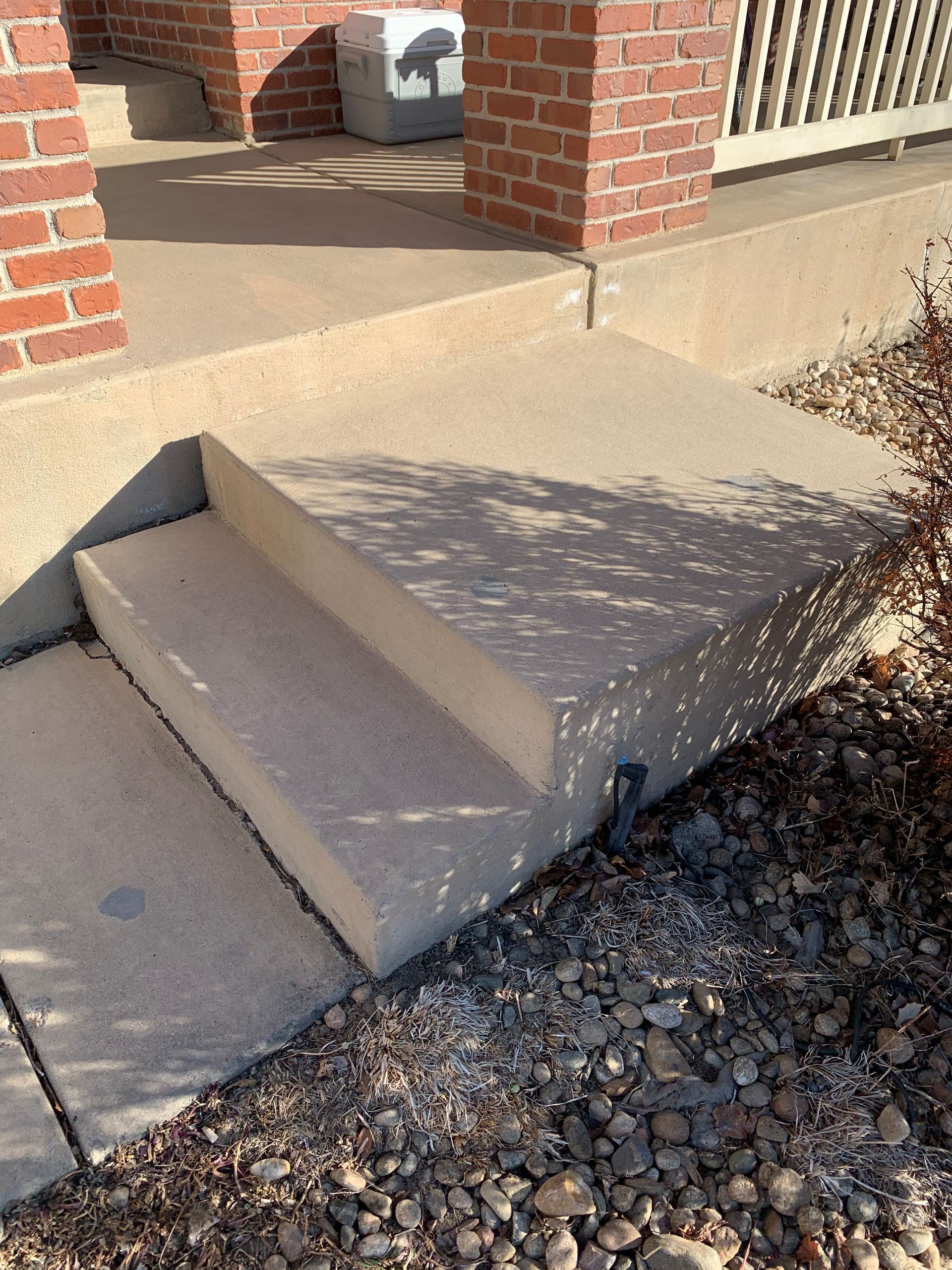 Concrete steps leading up to a porch with a brick pillar, gravel and dry leaves around the base.