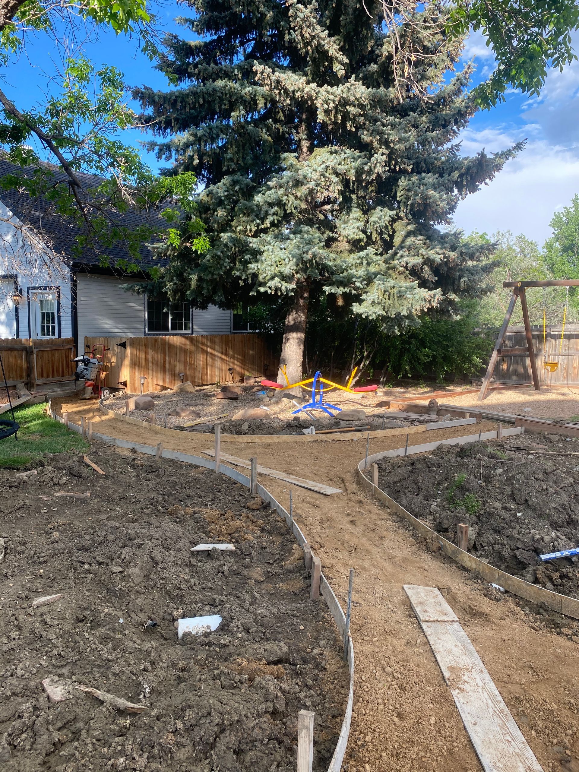 A backyard with a newly constructed gravel path, fenced area, large tree, and a swing set.