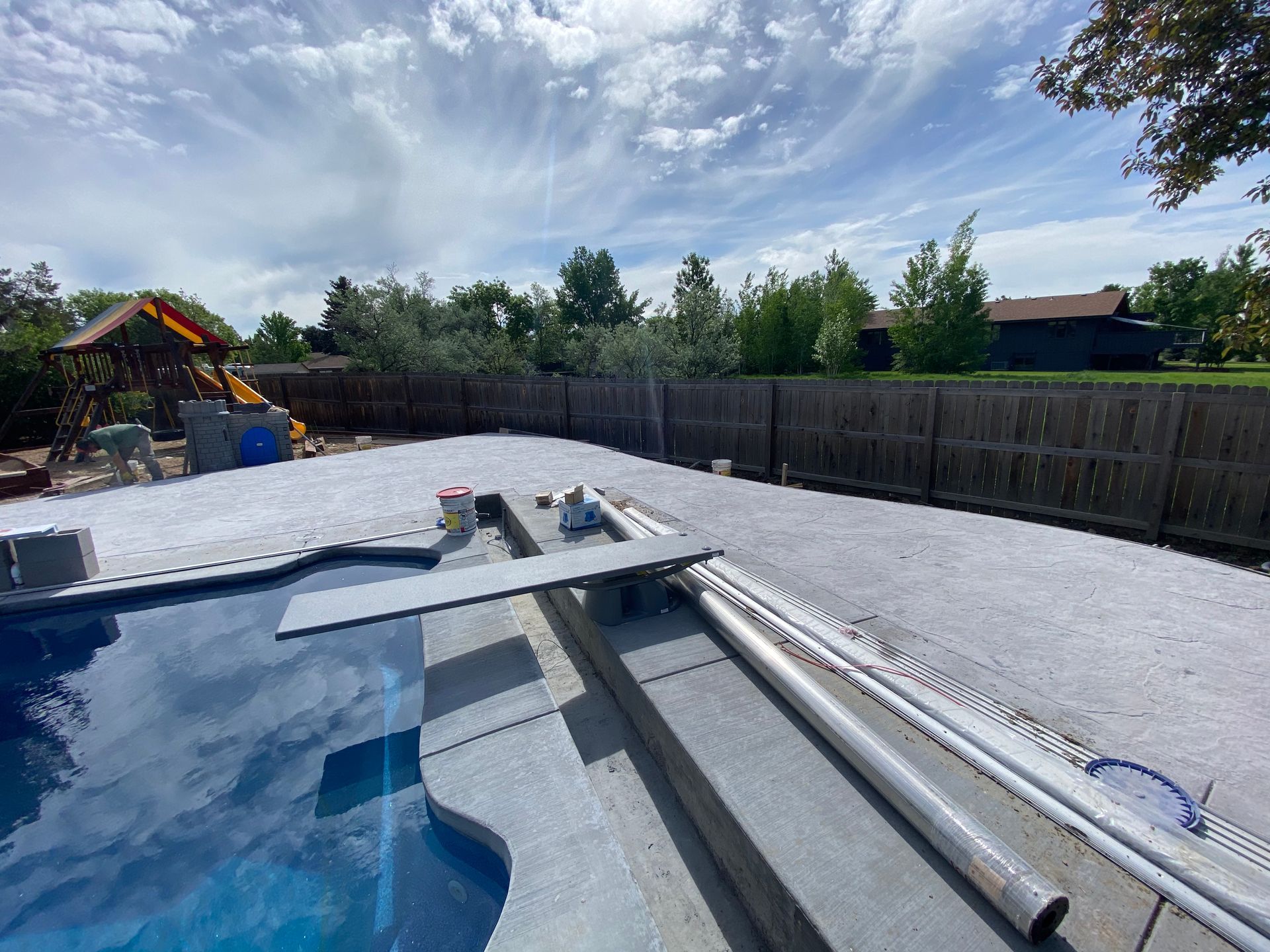 Backyard with a pool, diving board, and gravel surface. Fence and trees in the background under a cloudy sky.