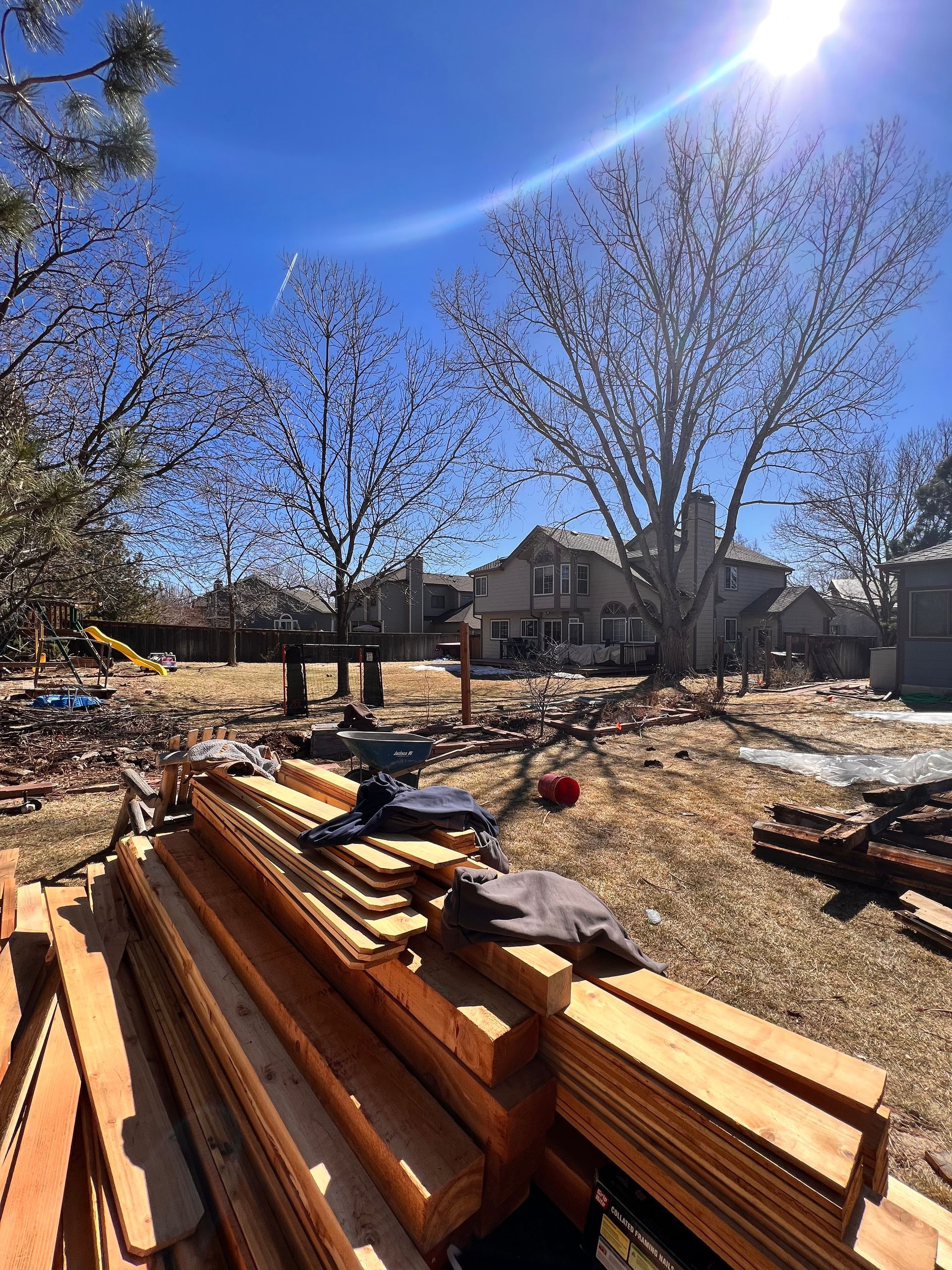 Backyard construction site; lumber pile in foreground, house and trees in background, sunny day.
