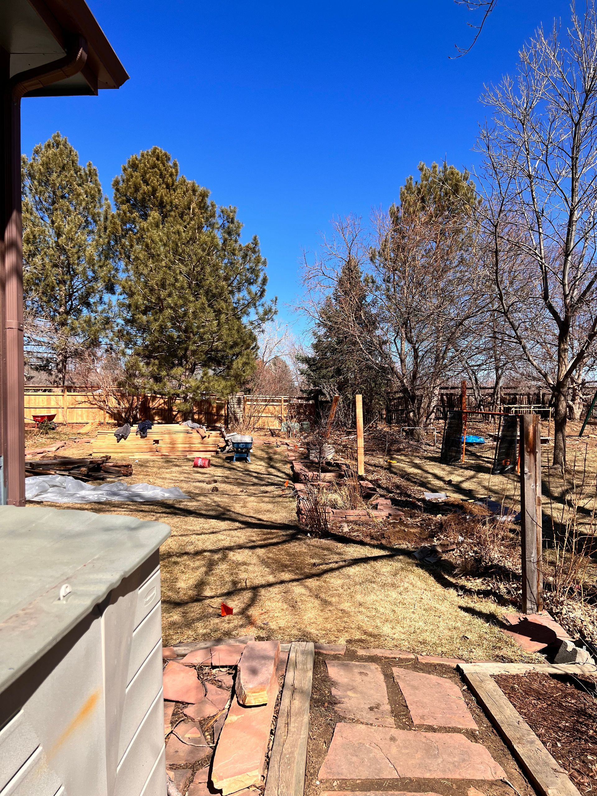 Backyard view, partially snow-covered, with trees, brick pathway, and bright blue sky.