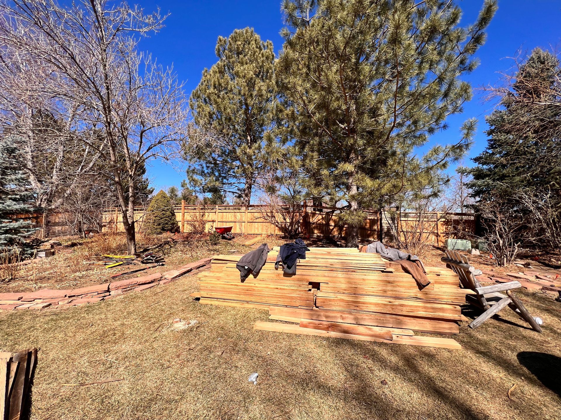 Backyard with wood planks, grass, trees, and fence under a bright blue sky.
