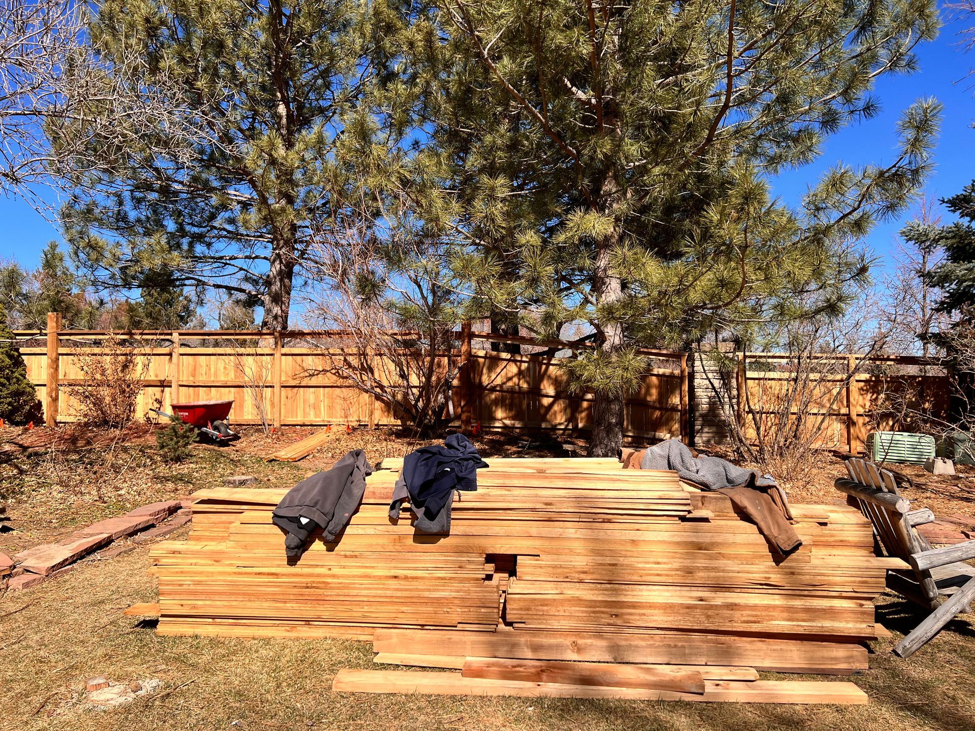 Pile of wooden planks in a yard with a brown fence and trees; three jackets lay on the wood.